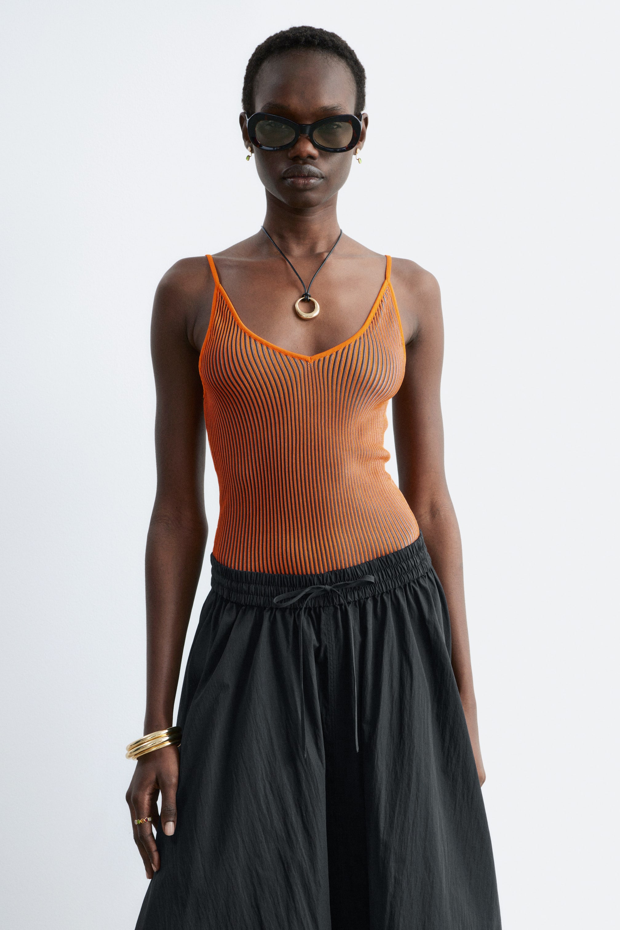 Agrandir l'image: Un débardeur côtelé à rayures orange et noires avec un profond décolleté en V et de fines bretelles est porté par le mannequin, qui porte également un pantalon noir à jambes larges.