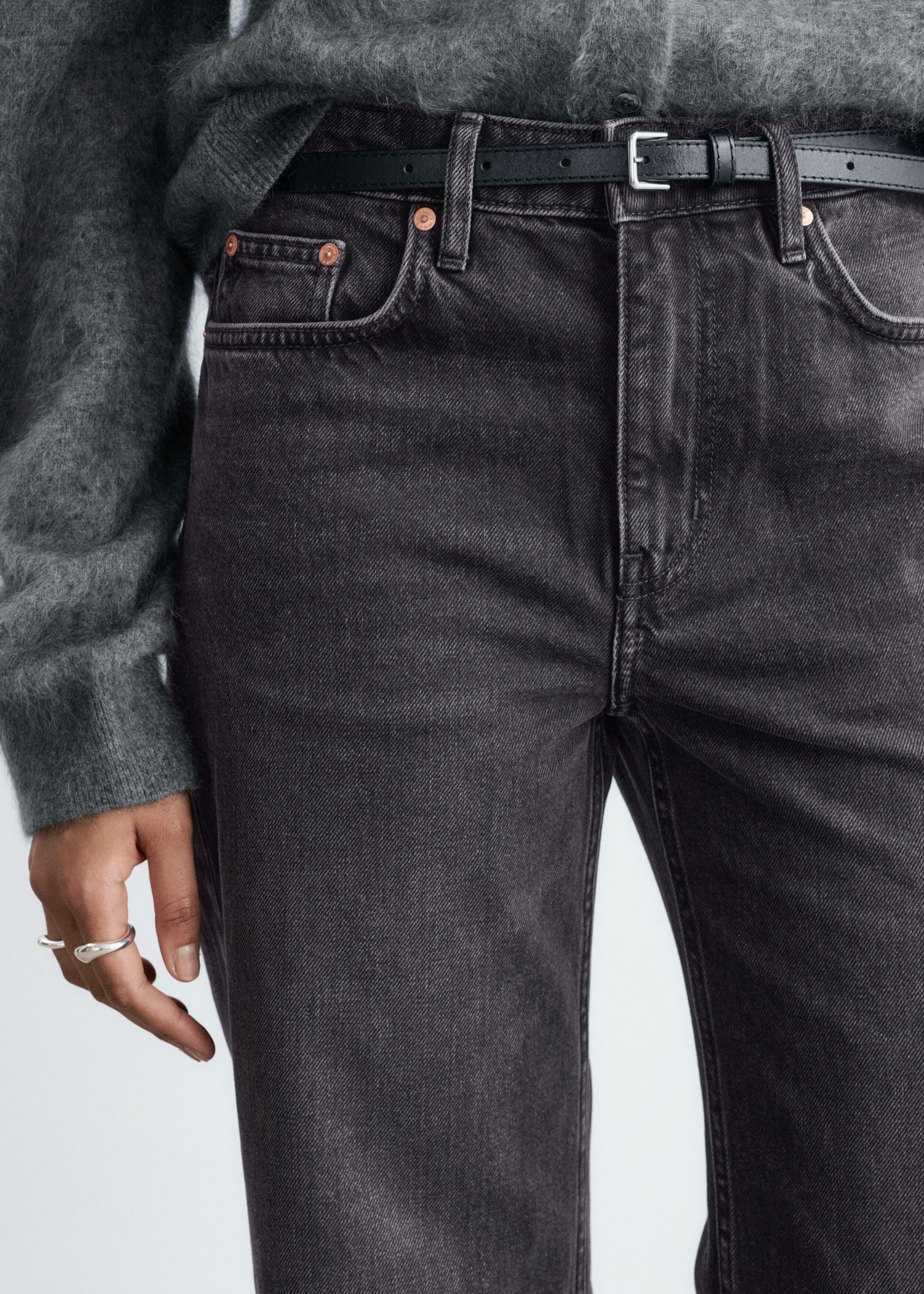 Dark gray washed denim jeans featuring a five-pocket design with copper rivets are being worn by the model. The jeans are styled with a thin black leather belt and a fuzzy gray knit sweater tucked into the waistband.
