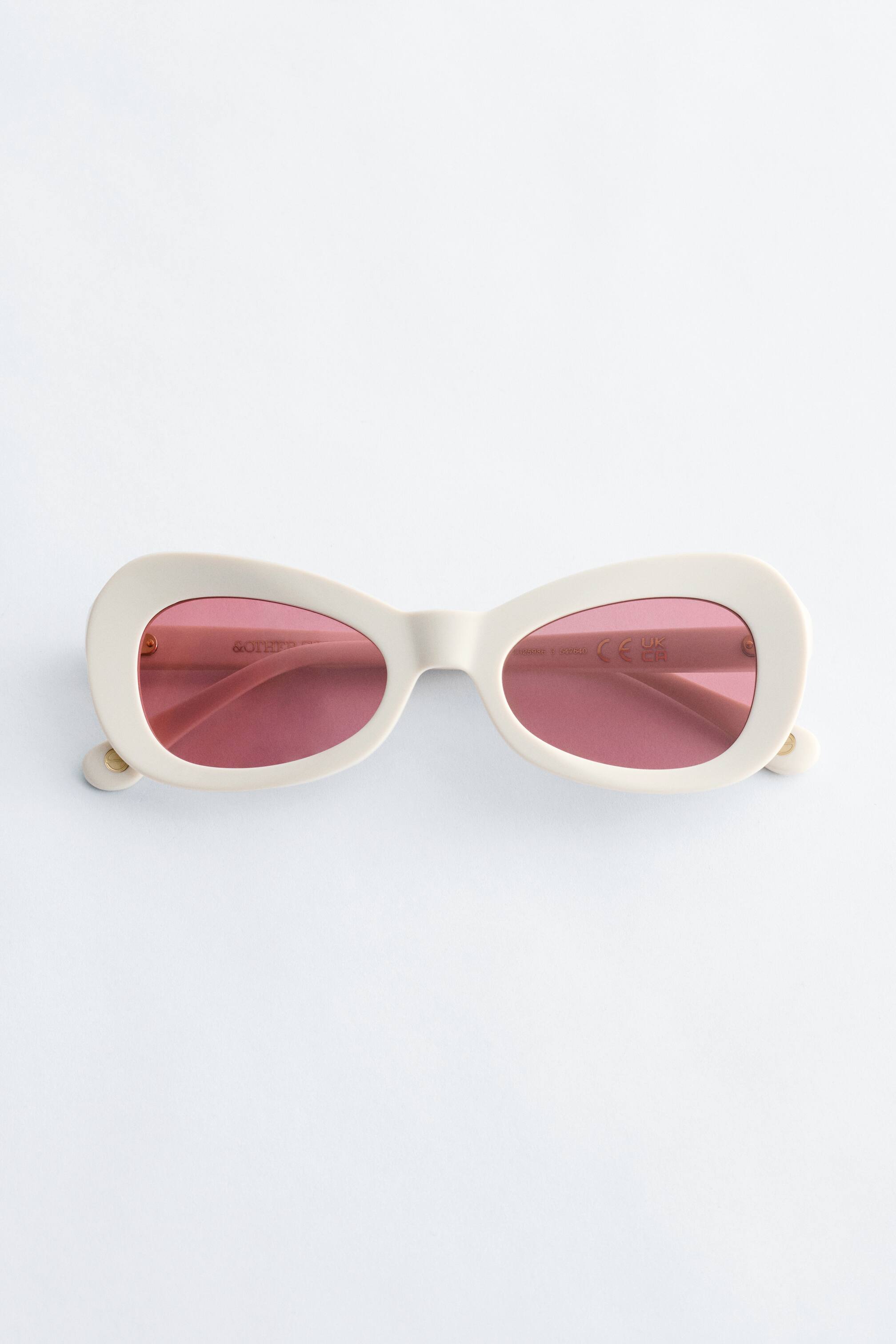 Προβολή μεγαλύτερης εικόνας: Τα γυαλιά ηλίου διαθέτουν σκελετούς από ανοιχτόχρωμη κρεμ ακετάτη, διαμορφωμένους σε ρετρό οβάλ σιλουέτα με μια διακριτική κίνηση γατίσιου ματιού στις εξωτερικές άκρες, και φακούς με ροζ απόχρωση. Παρουσιάζονται από μπροστινή όψη.