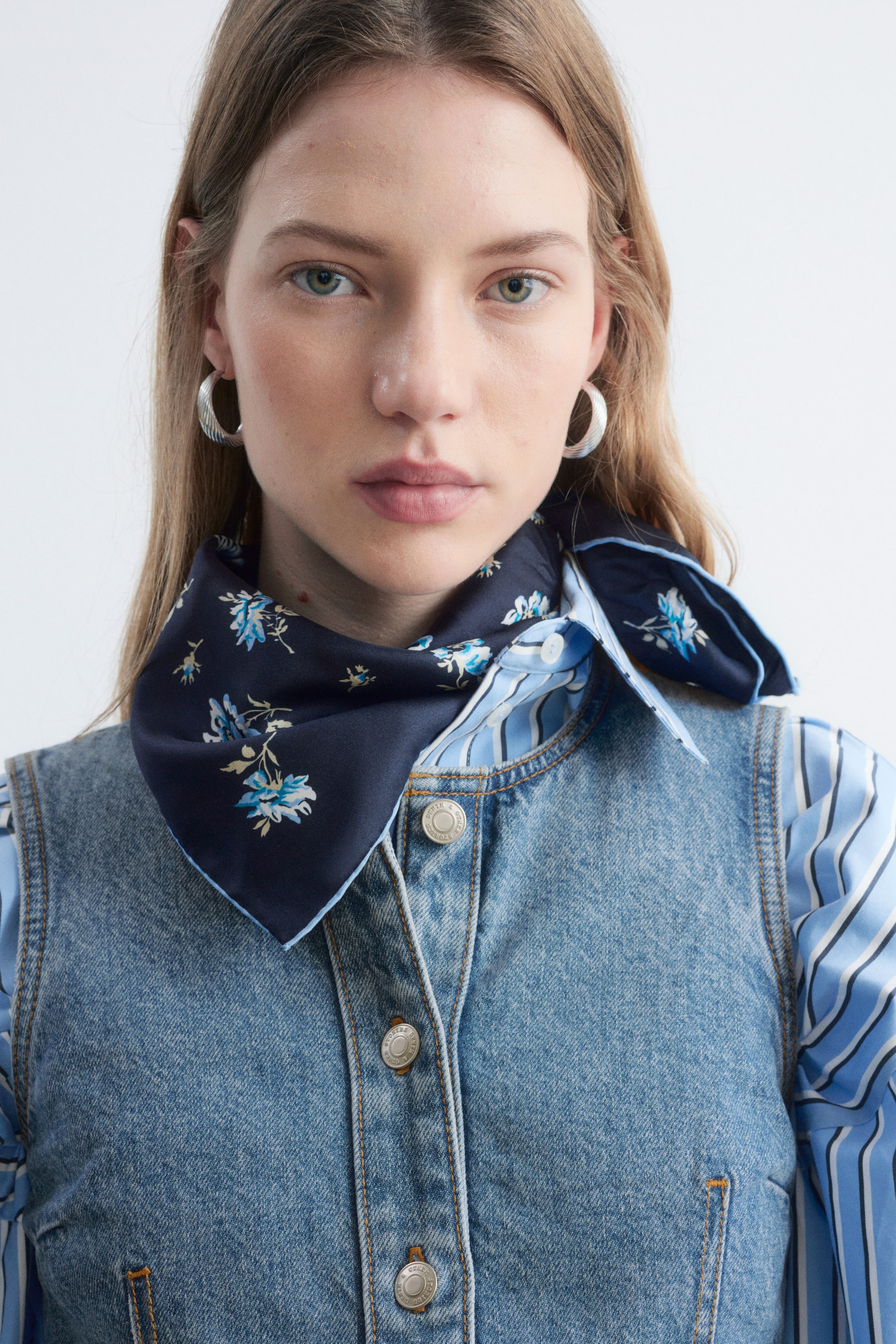 Größeres Bild anzeigen: Ein quadratisches Seidentuch in Dunkelblau mit hellblauem Blumenprint und Rand wird vom Model getragen. Es ist um den Hals gebunden und über einem gestreiften Hemd sowie unter einer Denim-Weste getragen.