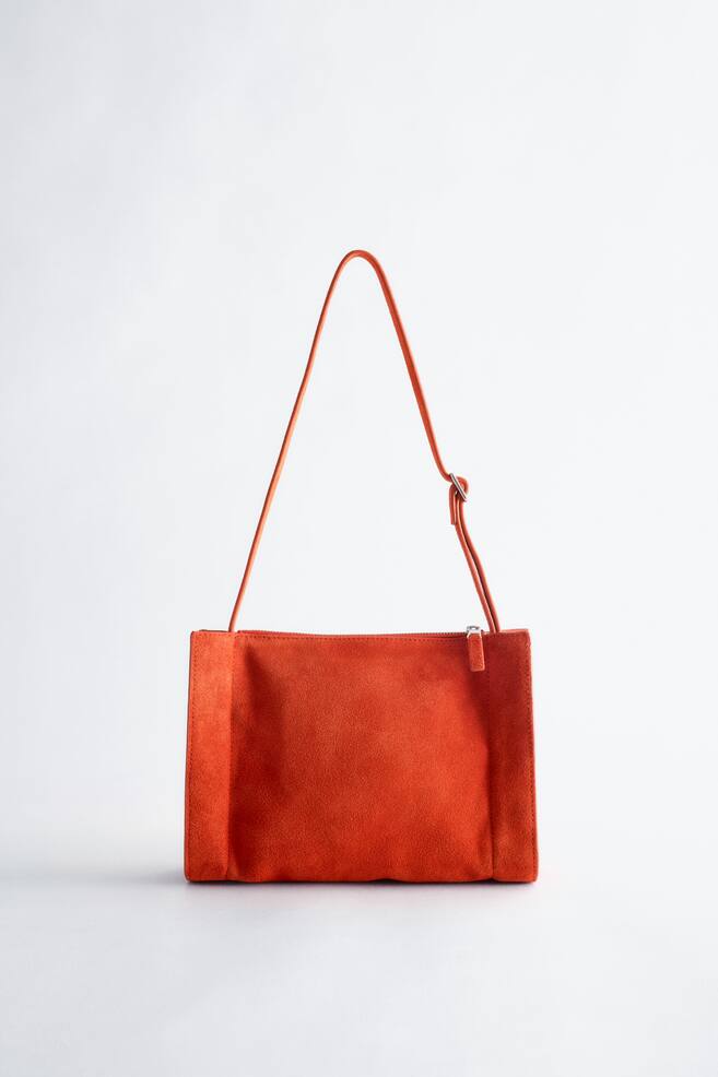 Le sac bandoulière en daim orange vif se présente de face, avec un corps rectangulaire et une fine sangle ajustable dotée d'une boucle argentée. Une fermeture éclair assortie est visible le long du bord supérieur du sac. - 1