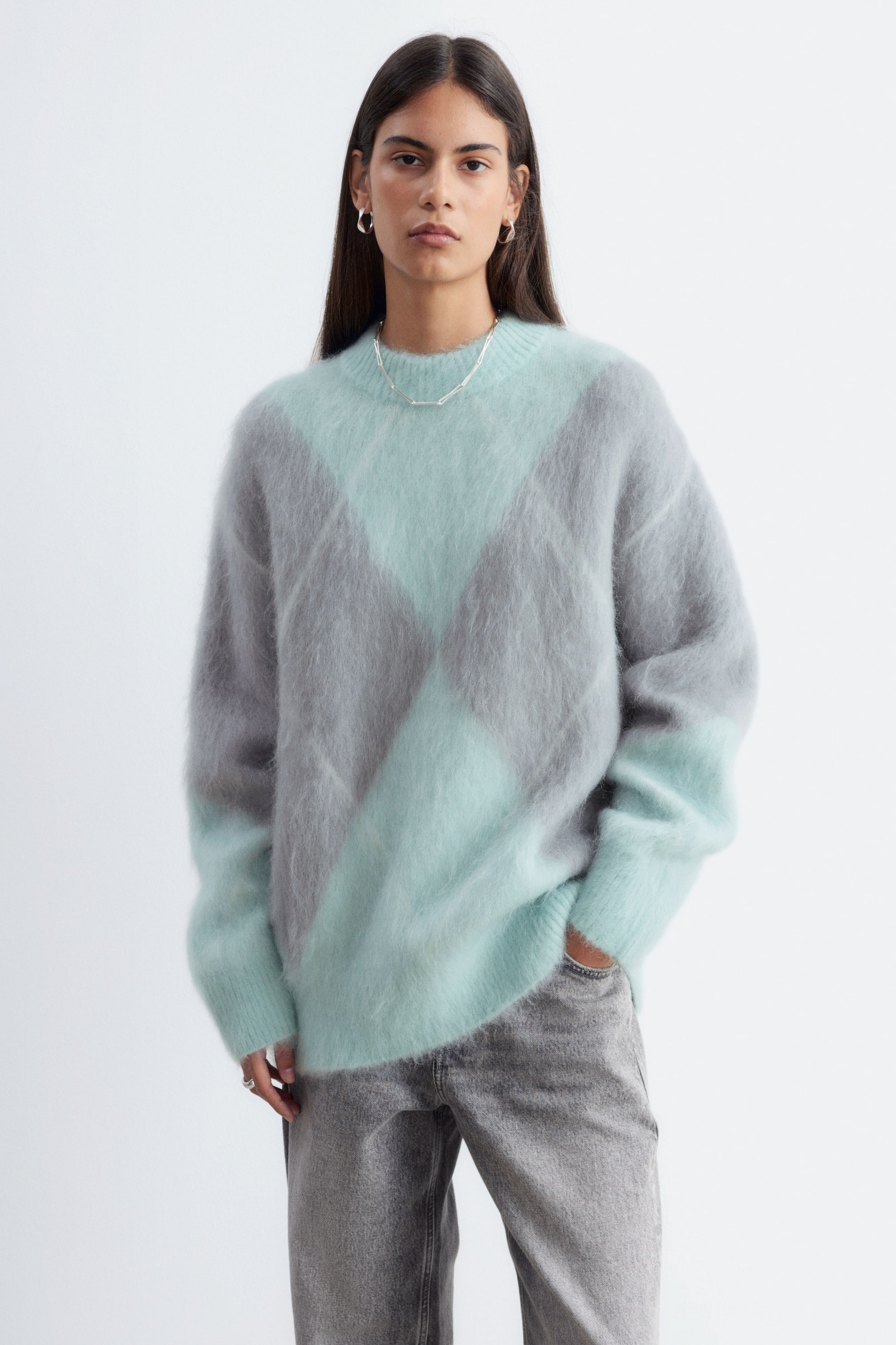 View larger image: An oversized mohair-blend jumper featuring a fuzzy texture and a mint green and grey diamond jacquard pattern is being worn by the model. The crew-neck knit is styled with grey denim jeans, with the model resting one hand in a pocket.