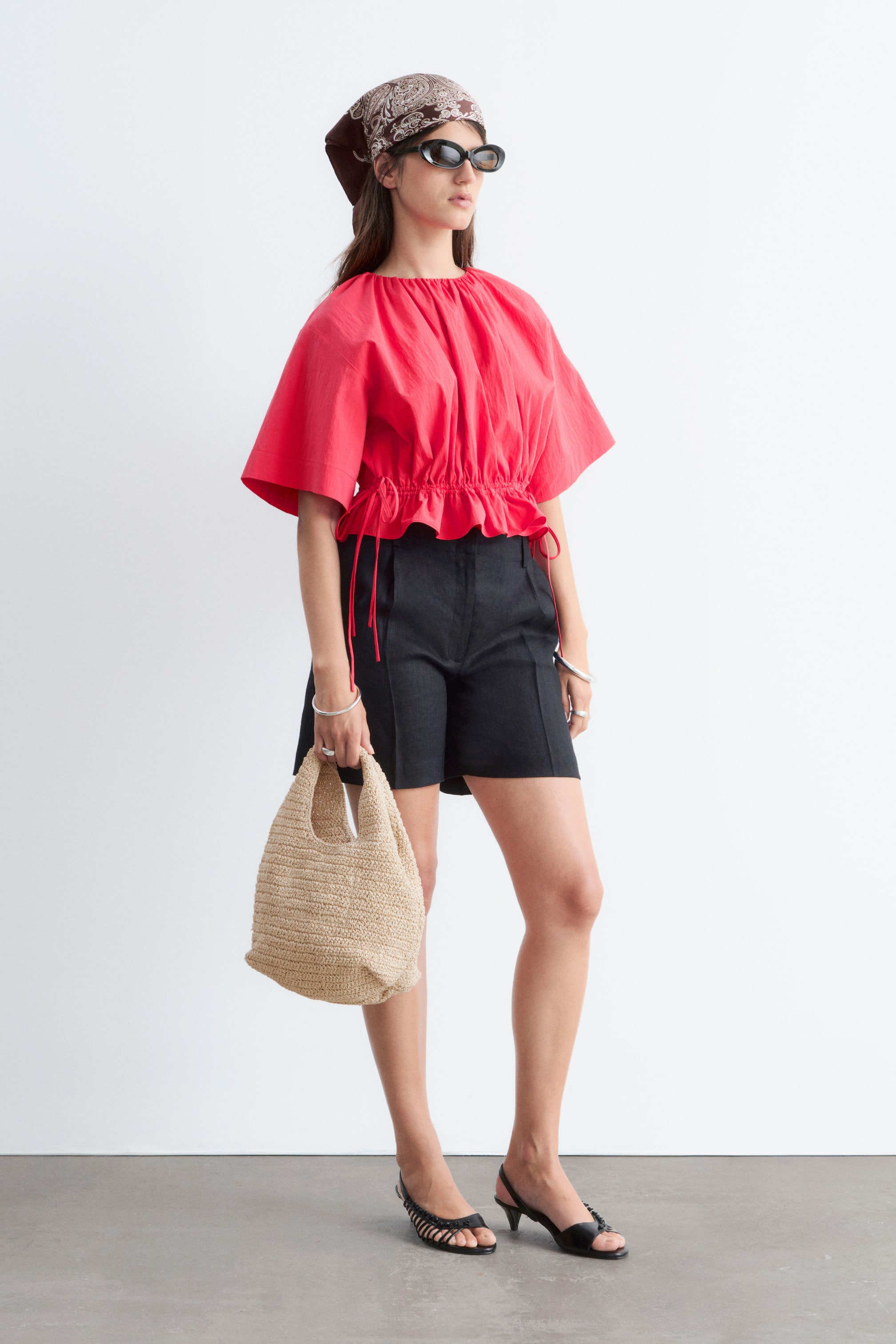 Zobrazit větší obrázek: Červený top má nařasený kulatý výstřih, objemné krátké rukávy a stahovací pas, který látku stáhne do nařaseného lemu, přičemž šňůrky volně visí přes černé šortky.