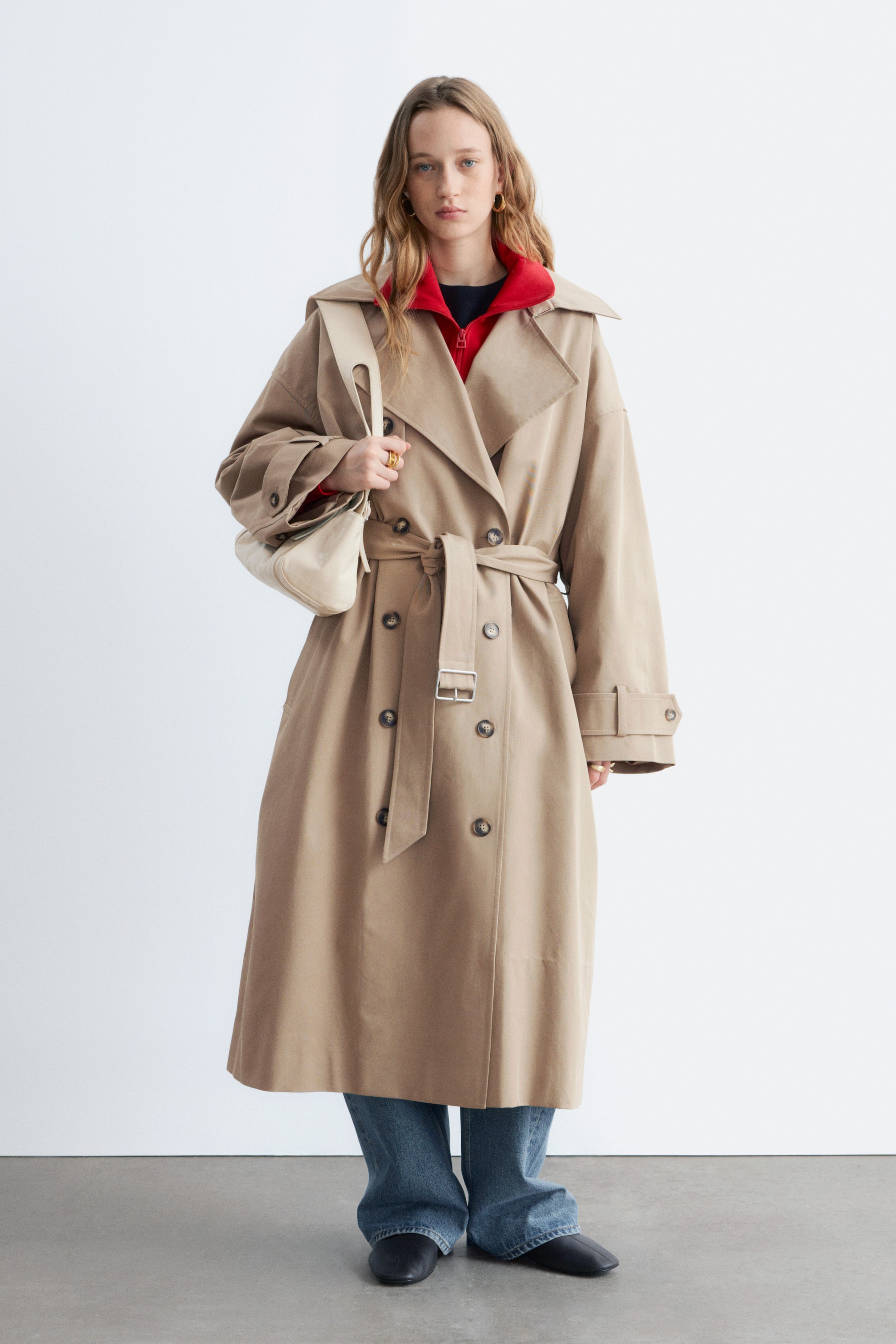 Ver imagen más grande: La modelo lleva un abrigo trench beige de doble botonadura, con el cinturón de tela atado a la cintura y las trabillas visibles en los puños. La modelo combina el abrigo con una blusa roja y lleva un bolso de mano de color claro.