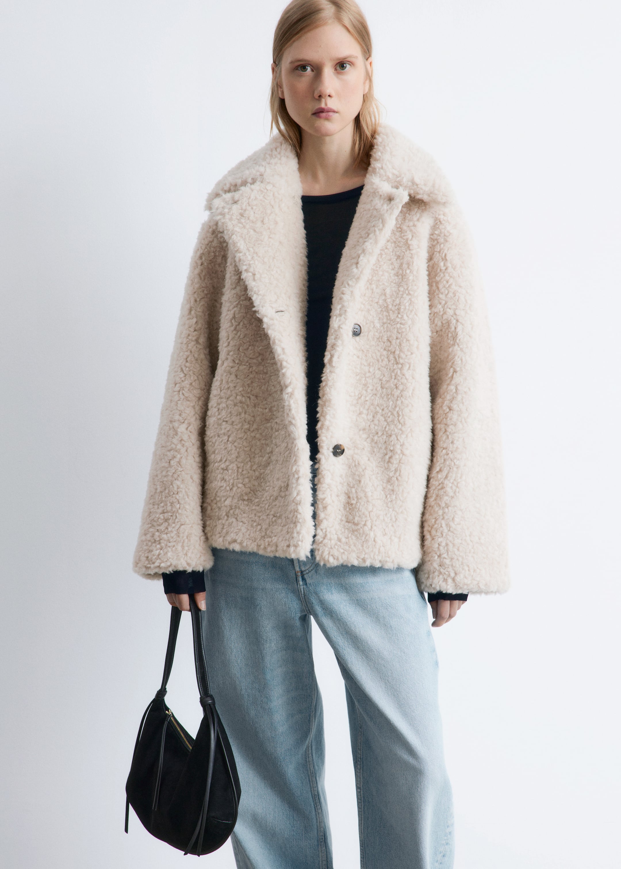 Se større billede: En beige teddyfrakke vender let mod højre og fremviser en bred, spredt krave og en afslappet, kort pasform. Den har to synlige brune knapper på frontlukningen, og en sort langærmet top er lagdelt under.