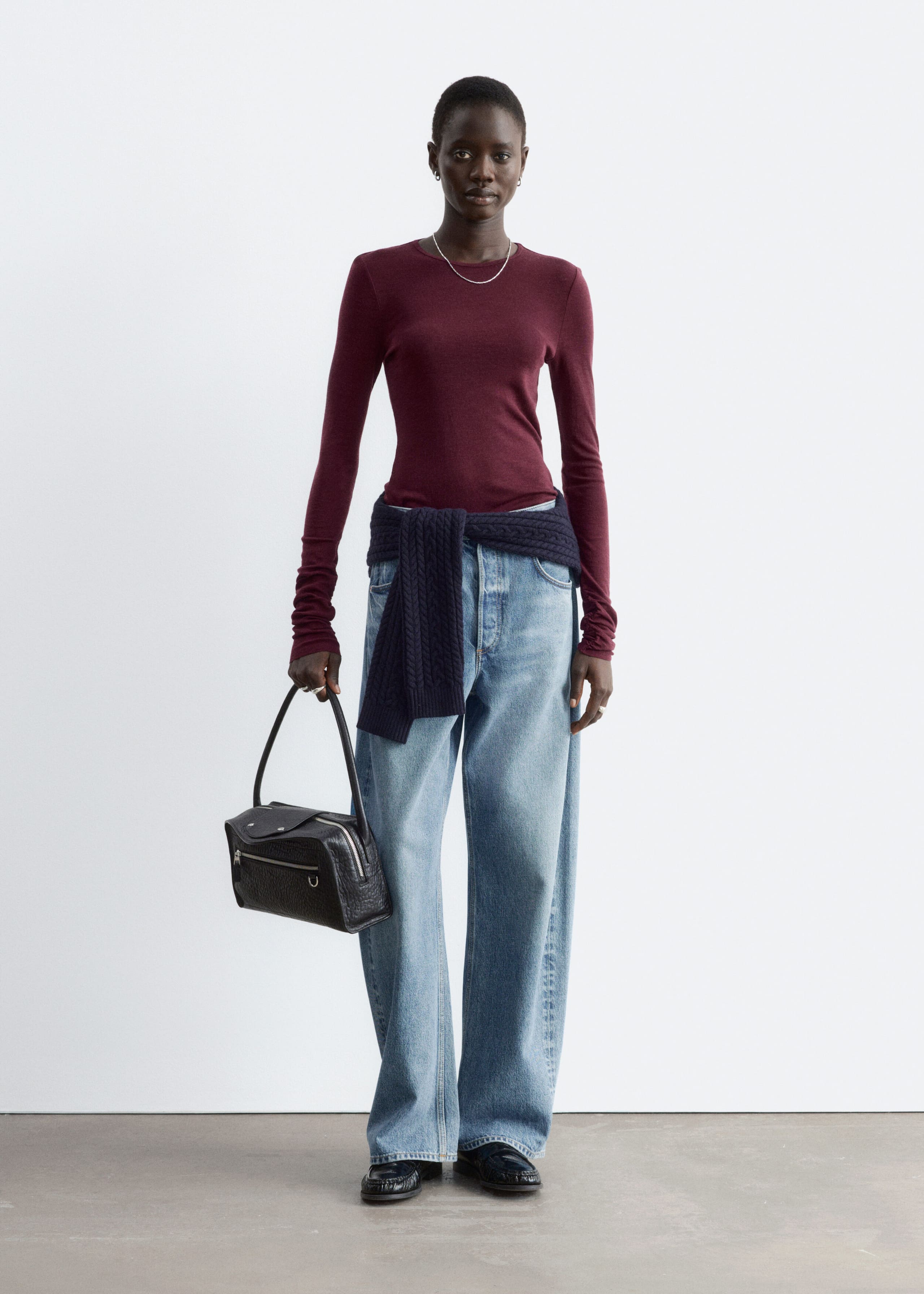 Ein schlicht gestricktes Wolloberteil in einem tiefen Burgunderrot wird vom Model getragen. Das langärmelige Oberteil mit Rundhalsausschnitt ist in eine helle Wide-Leg-Jeans gesteckt, dazu ist ein dunkelblauer Strickpullover um die Taille gebunden.