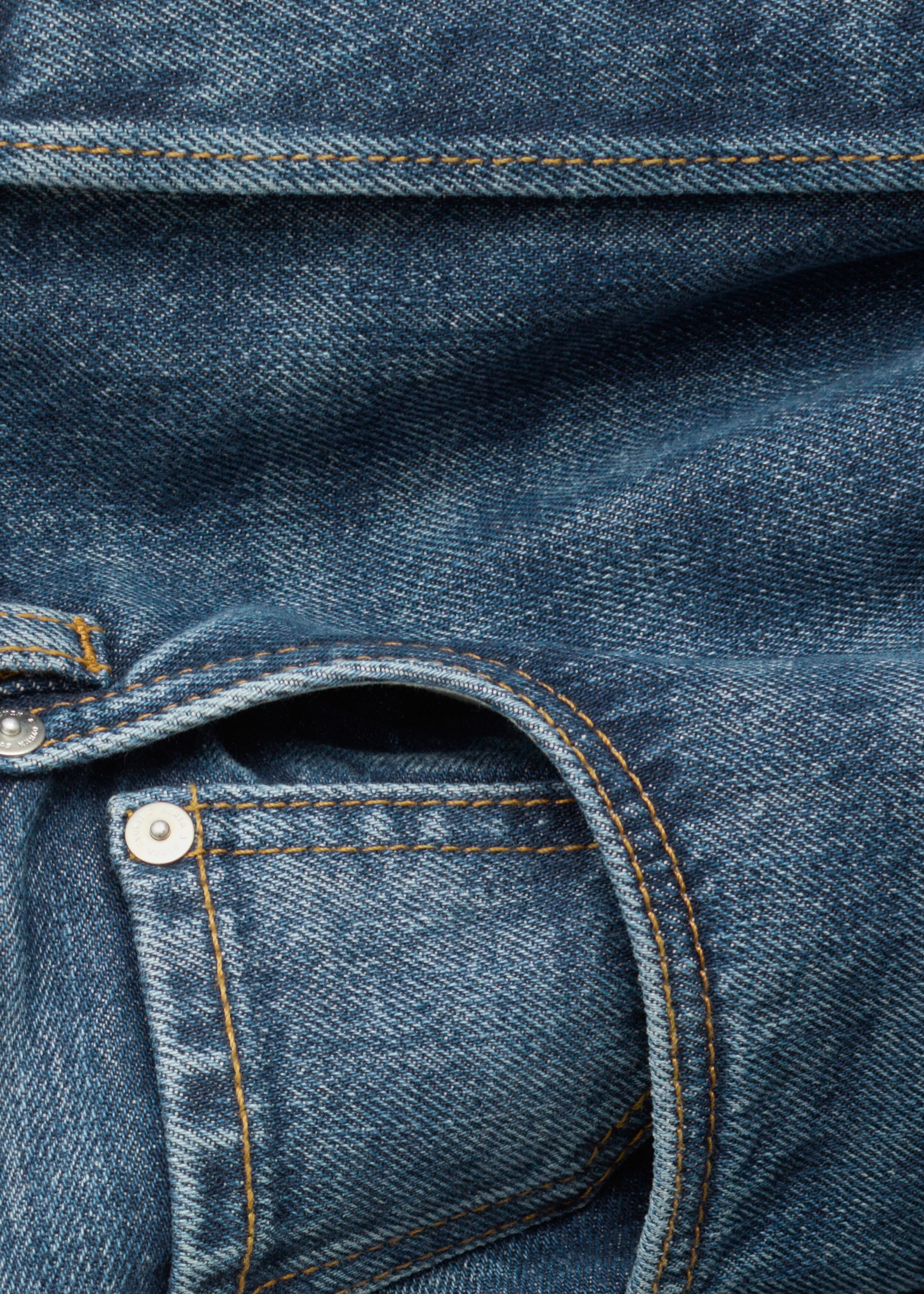 A close-up view shows the front pocket of blue denim jeans, highlighting the curved main pocket and a small coin pocket. The pockets feature contrasting orange topstitching and a silver metal rivet on the textured denim fabric.