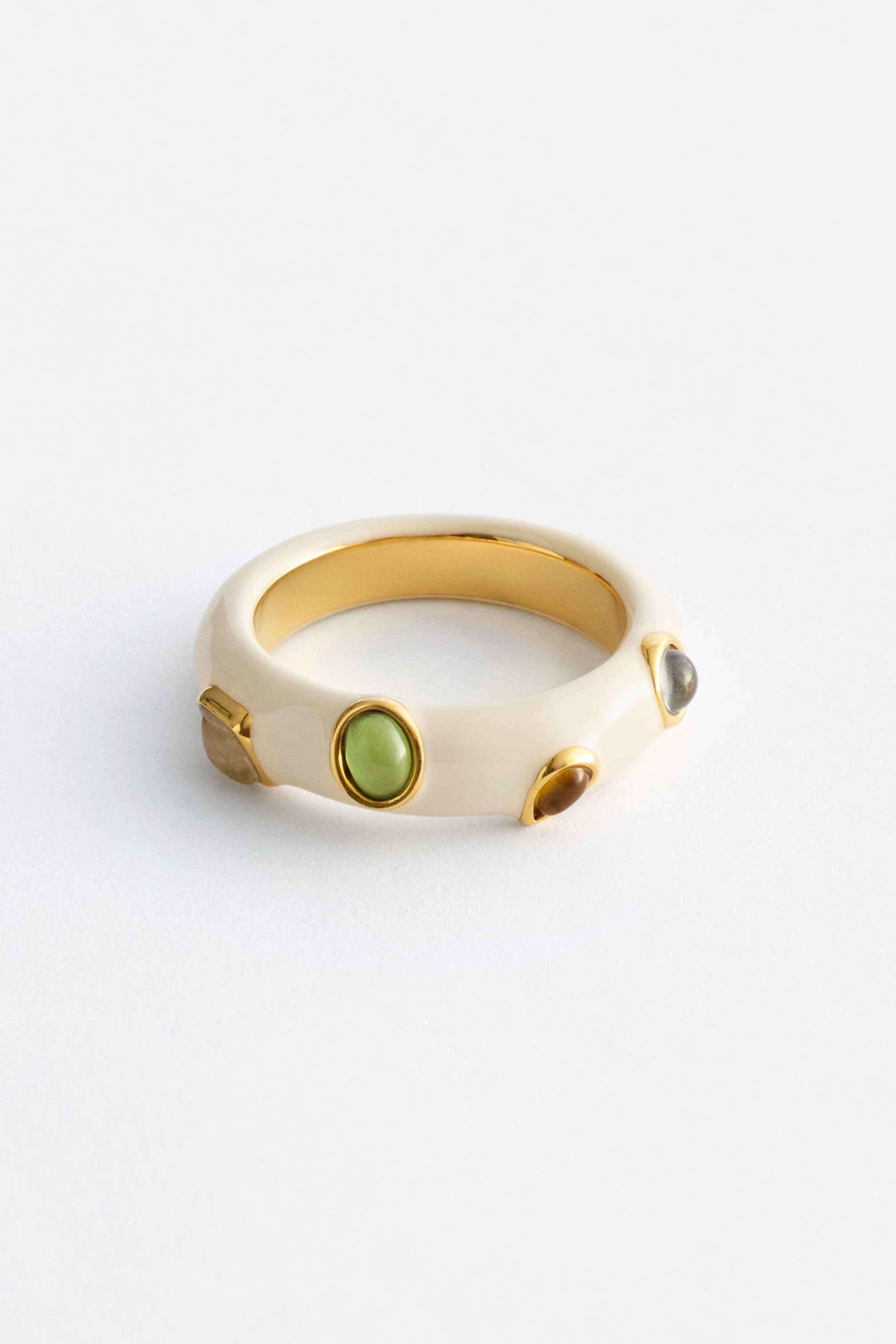 Größeres Bild ansehen: Ein breiter Ring mit einem elfenbeinfarbenen Emaille-Außenbereich und einem polierten goldfarbenen Innenbereich, der verschiedene Cabochon-Steine in unterschiedlichen Formen und Farben präsentiert, darunter ein runder grüner, ein ovaler brauner und ein runder grauer Stein, alle in goldfarbenen Fassungen gefasst.