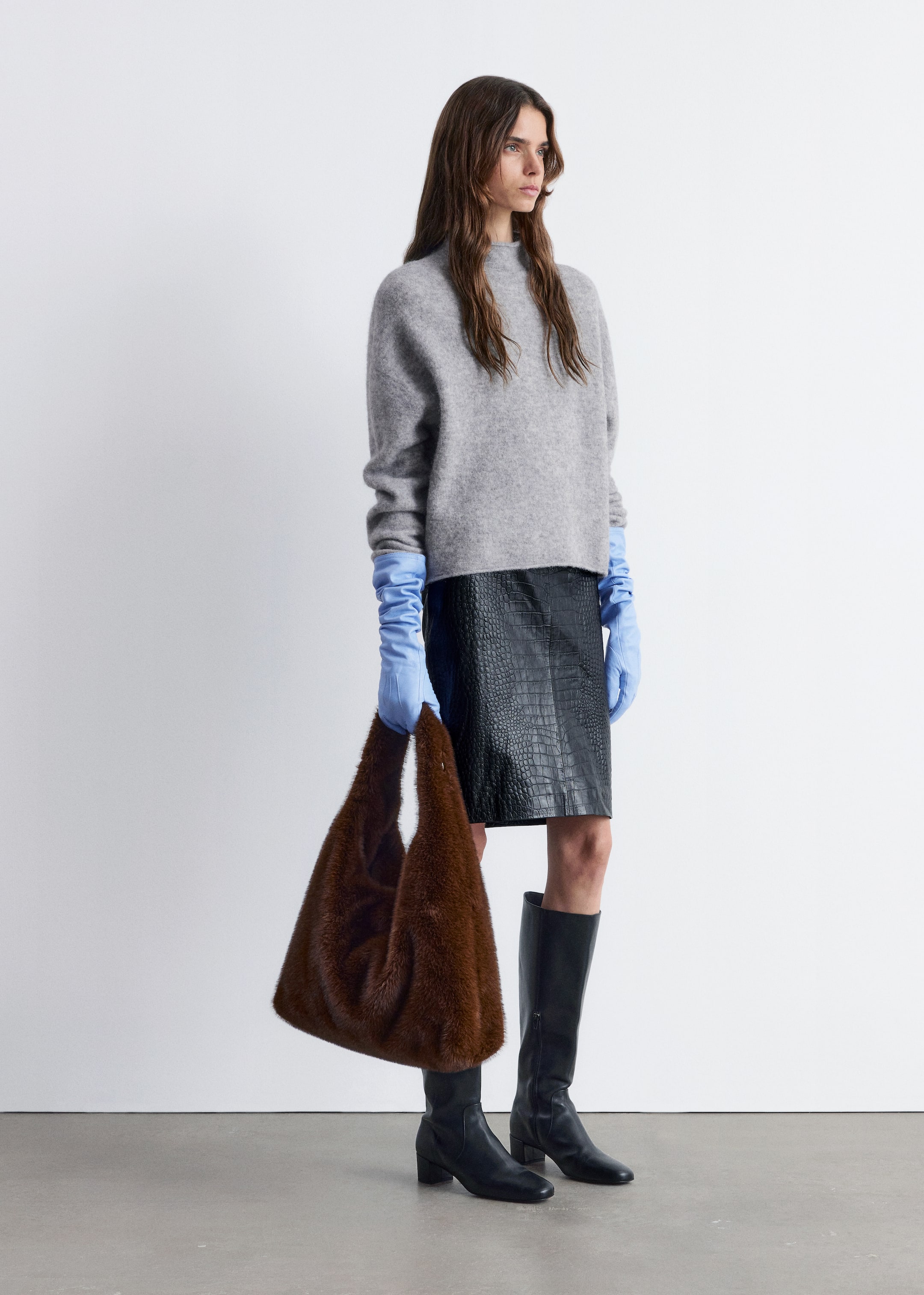 Visa större bild: En modell presenterar en outfit med en grå polotröja med lös passform, en svart minikjol i falsk krokodil och svarta knähöga stövlar. Ensemblen är accessoriserad med ljusblå långa handskar, skrynklade på armarna, och en brun lurvig slouchy väska som hålls i handen.