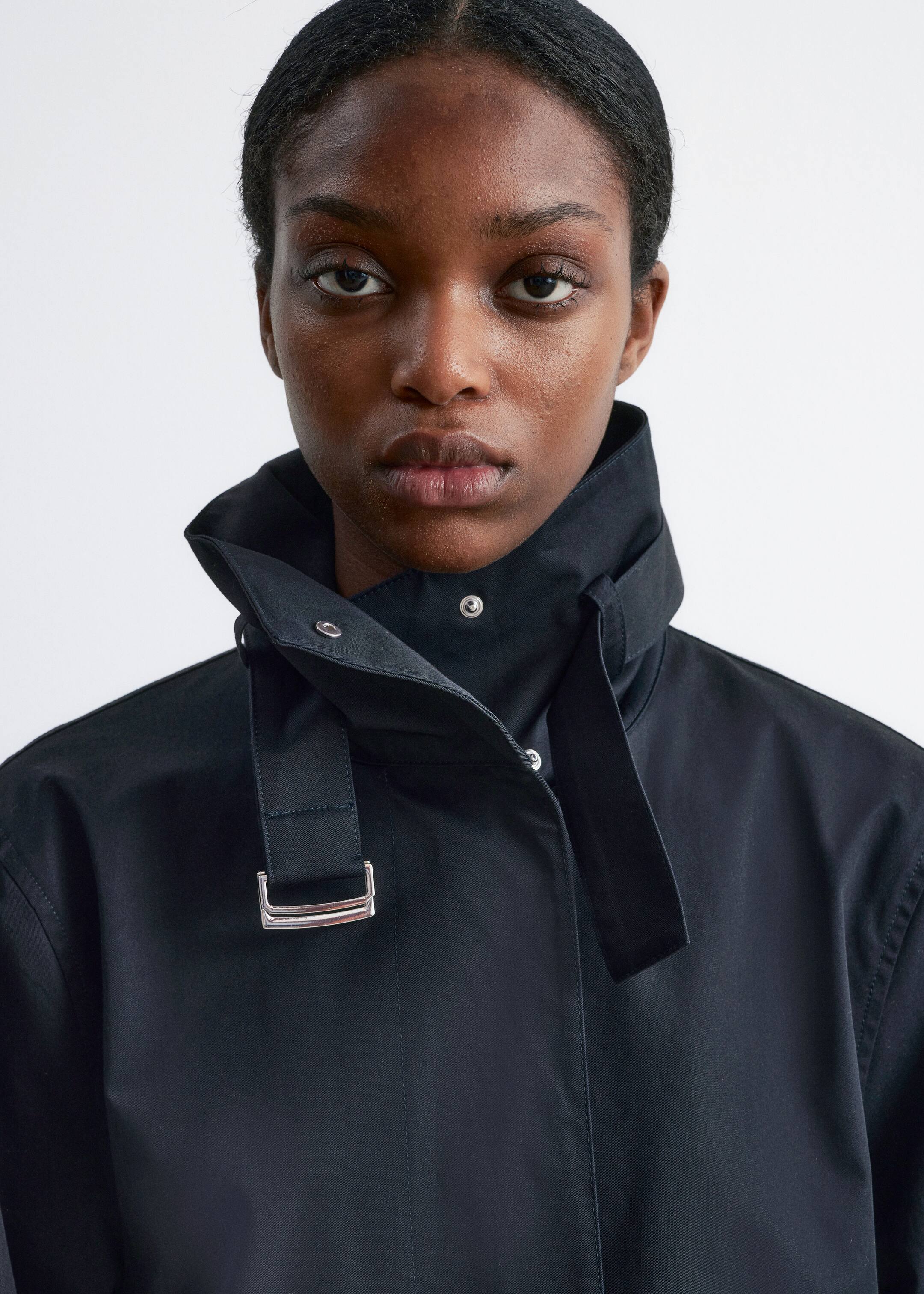Grösseres Bild ansehen: Eine schwarze Jacke wird aus einem frontalen Winkel präsentiert, mit einem hohen Stehkragen und sichtbaren silbernen Druckknöpfen. Ein breiter schwarzer Riemen mit einer silbernen rechteckigen Schnalle ist auffällig auf der oberen Vorderseite zu sehen und erstreckt sich nach unten.