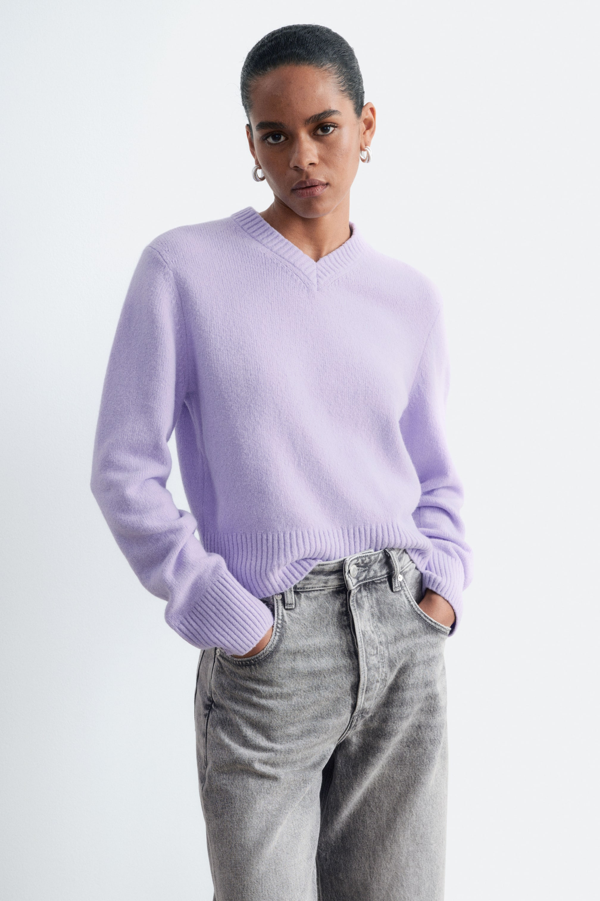 Visualizza l'immagine ingrandita: Un pullover lavorato a maglia color lilla con scollo a V a coste, ampi polsini a coste e fondo a coste è indossato dalla modella. Il pullover è abbinato a jeans grigi, con la modella che riposa le mani nelle tasche anteriori.