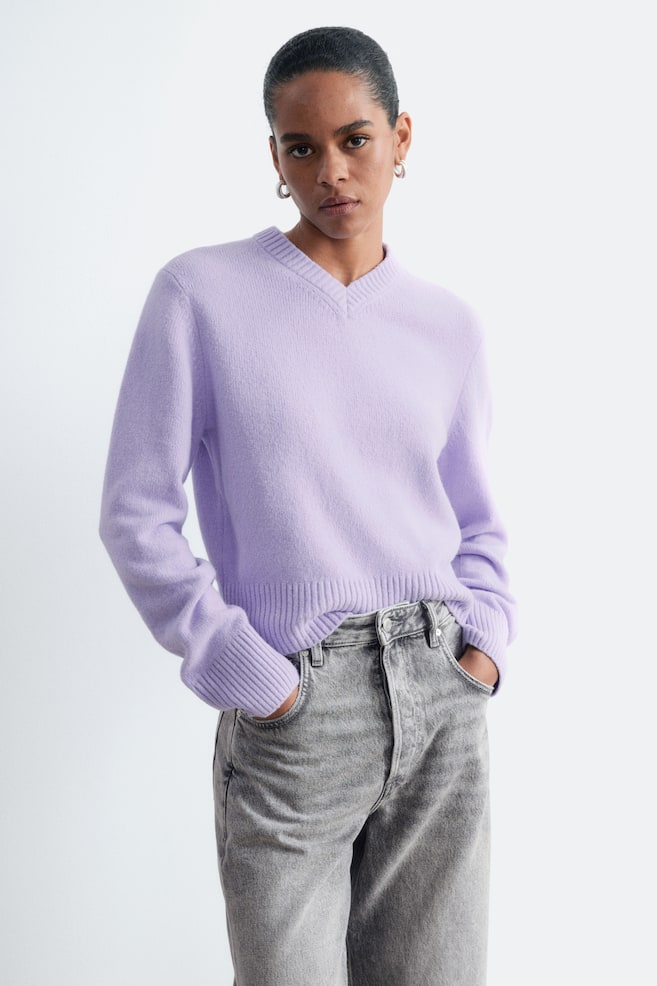 La modella indossa un maglione lavorato a maglia color lilla, caratterizzato da scollo a V a costine, ampi polsini a costine e orlo a costine. Il maglione è abbinato a jeans grigi e la modella tiene le mani nelle tasche anteriori. - 1