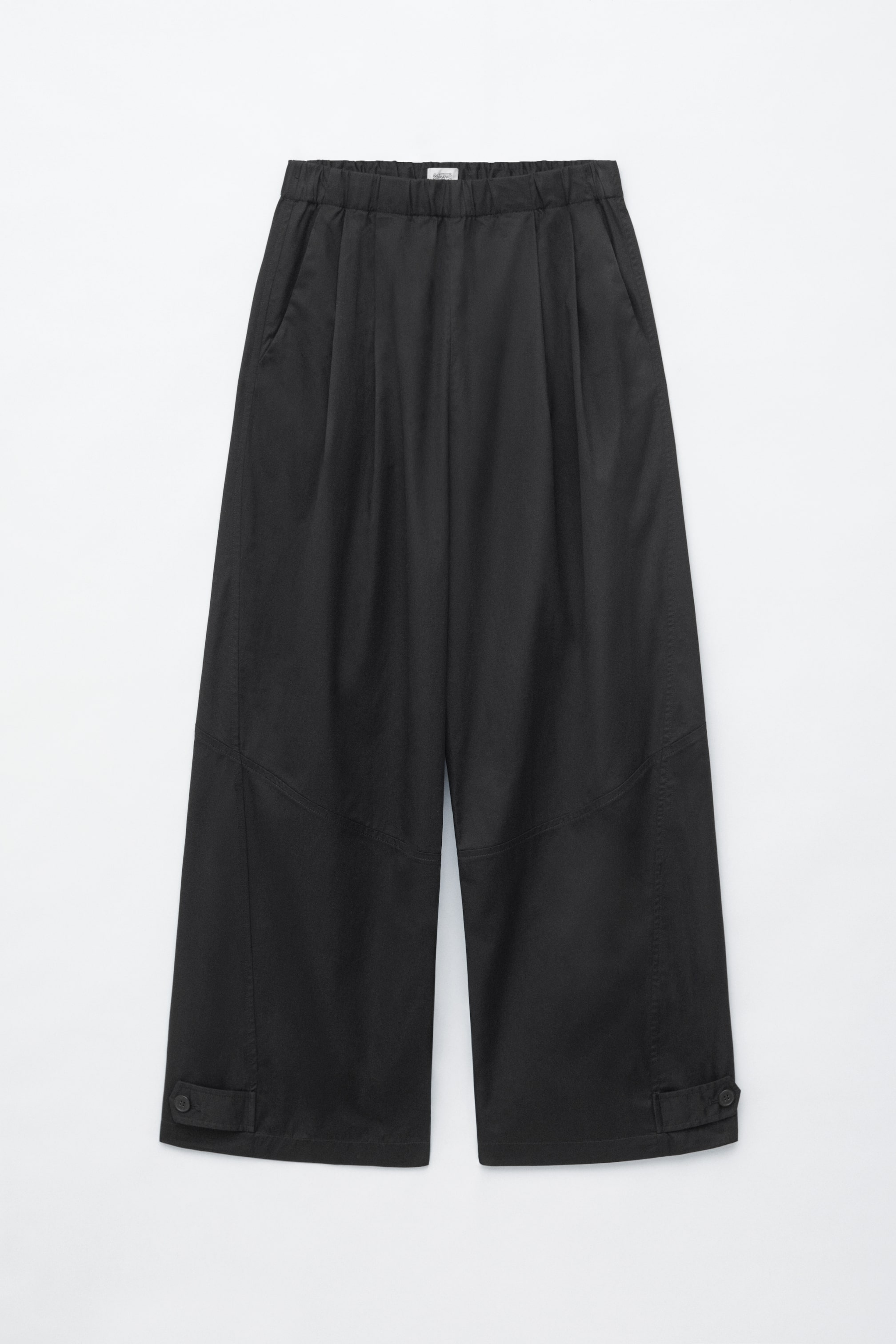 Grotere afbeelding bekijken: Zwarte broek met wijde pijpen, getoond in een vooraanzicht, met een elastische tailleband, vaste plooien aan de voorkant en zijnaadzakken. De broek toont gedetailleerde snijnaden op de onderbenen en verstelbare knoopsluitingen aan elke zoom.