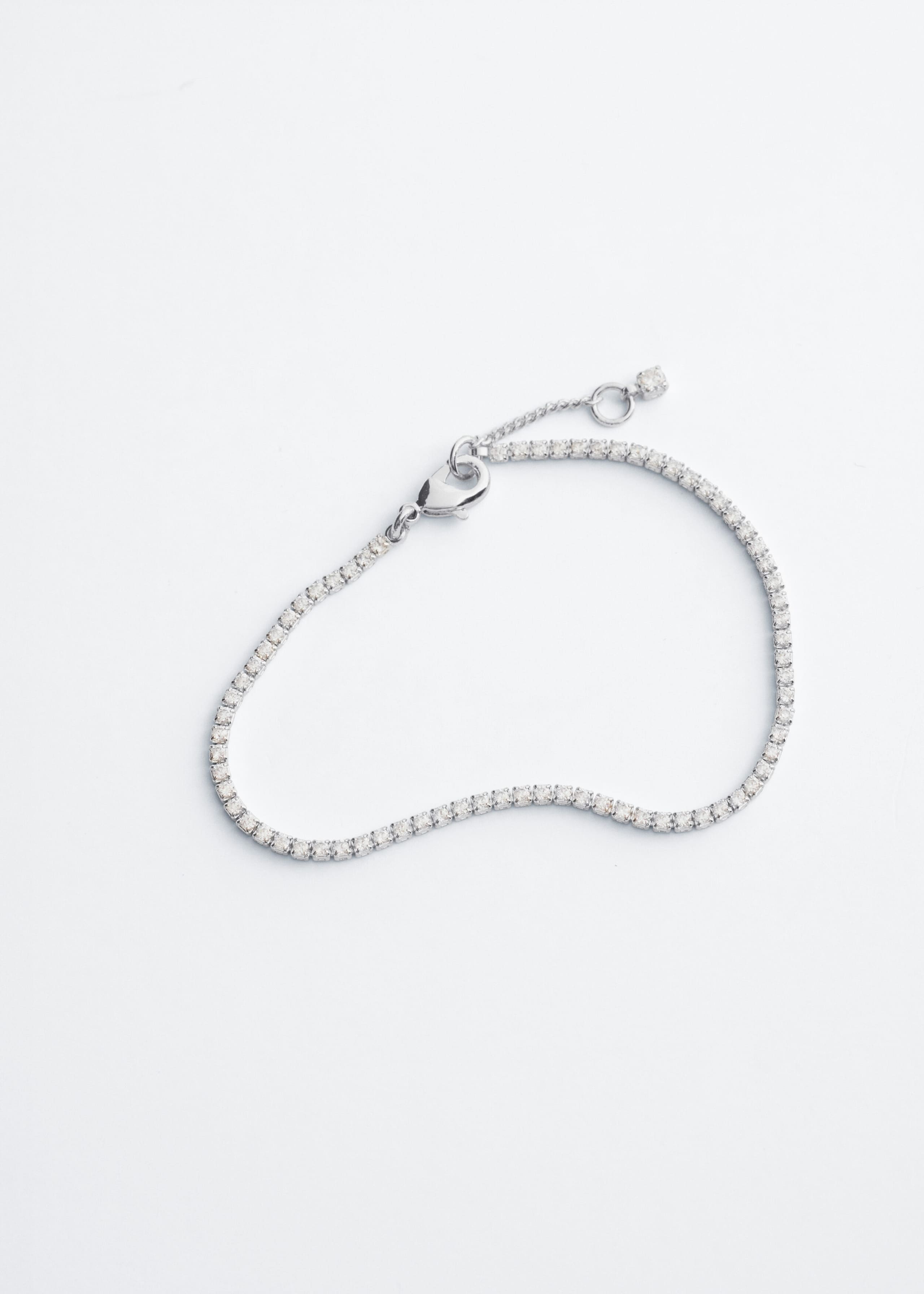 This silver-toned brass tennis bracelet lies flat in a top view, displaying a continuous row of small, glistening rhinestones. It features a lobster clasp closure and a short extension chain with a single rhinestone detail at the end.