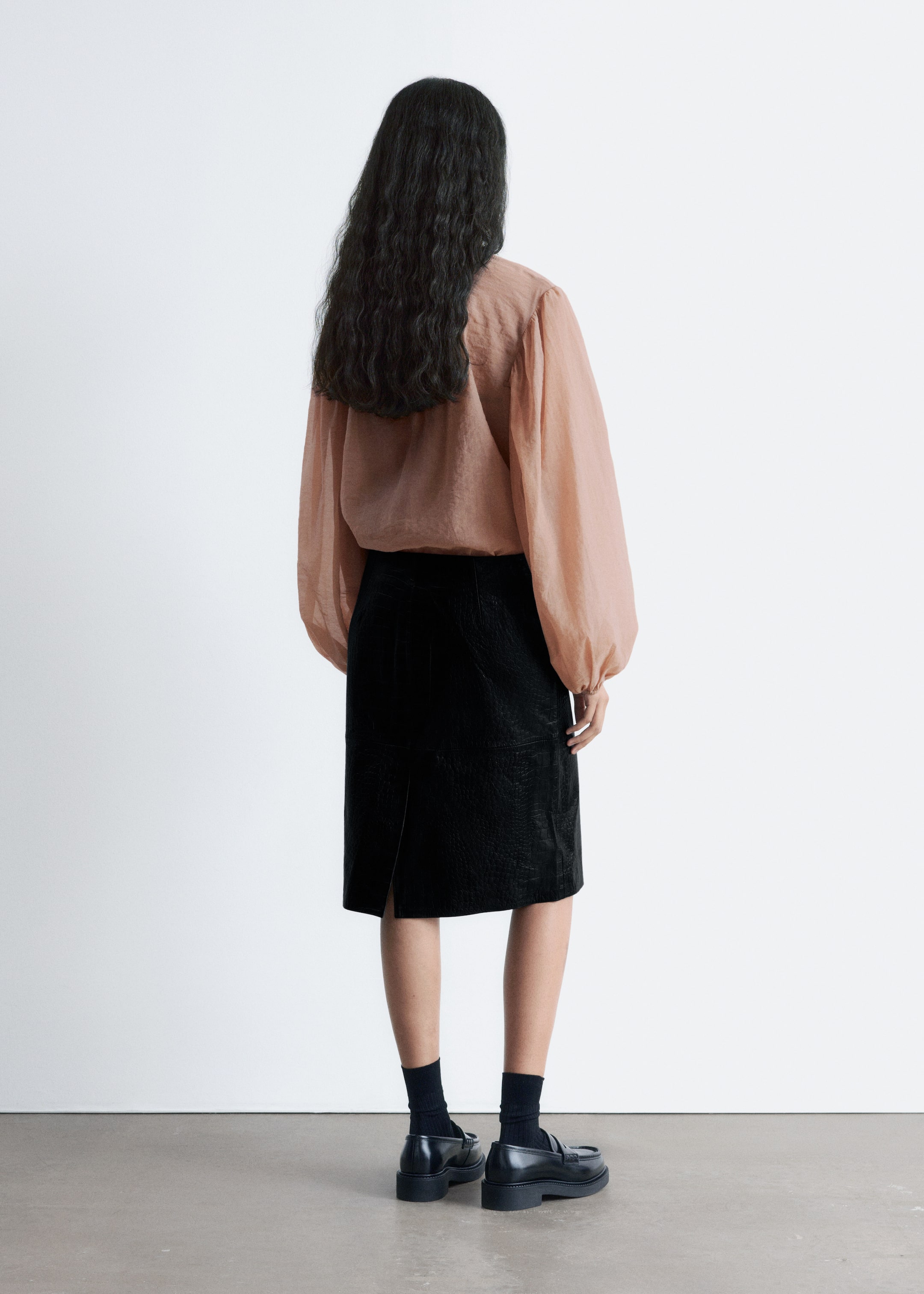 Grotere afbeelding bekijken: Het product, een lichtbruin-roze volumineuze blouse en een zwarte knielange rok, wordt van de achterkant gepresenteerd. De blouse heeft lange, geplooide ballonmouwen en een losse, vloeiende pasvorm, die in de rok lijkt te zijn gestopt, welke een subtiele krokodillen-embossed textuur en een achterste split toont.