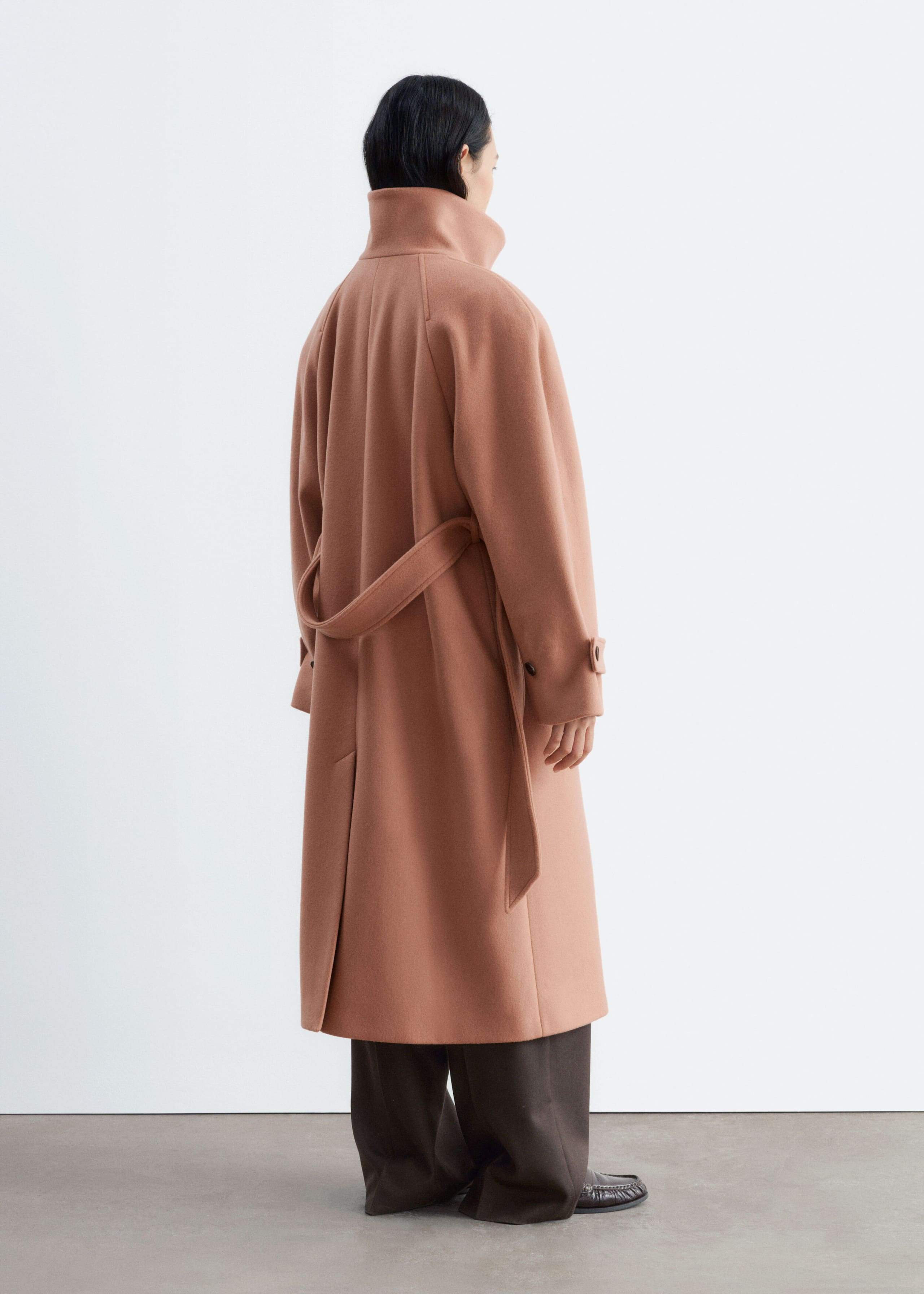 A dusty rose wool-blend coat is being worn by the model, shown from a three-quarter rear view. The coat features a high stand-up collar, a matching tie belt at the waist, and a back vent, and is styled with dark wide-leg trousers and shoes.