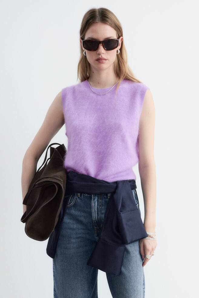 A light purple, sleeveless knitted vest in brushed cashmere is being worn by the model, showcasing its crew neckline and fuzzy texture. The vest is styled with blue jeans, a dark blue jumper tied around the waist, and a brown suede handbag being carried under the arm. - 1