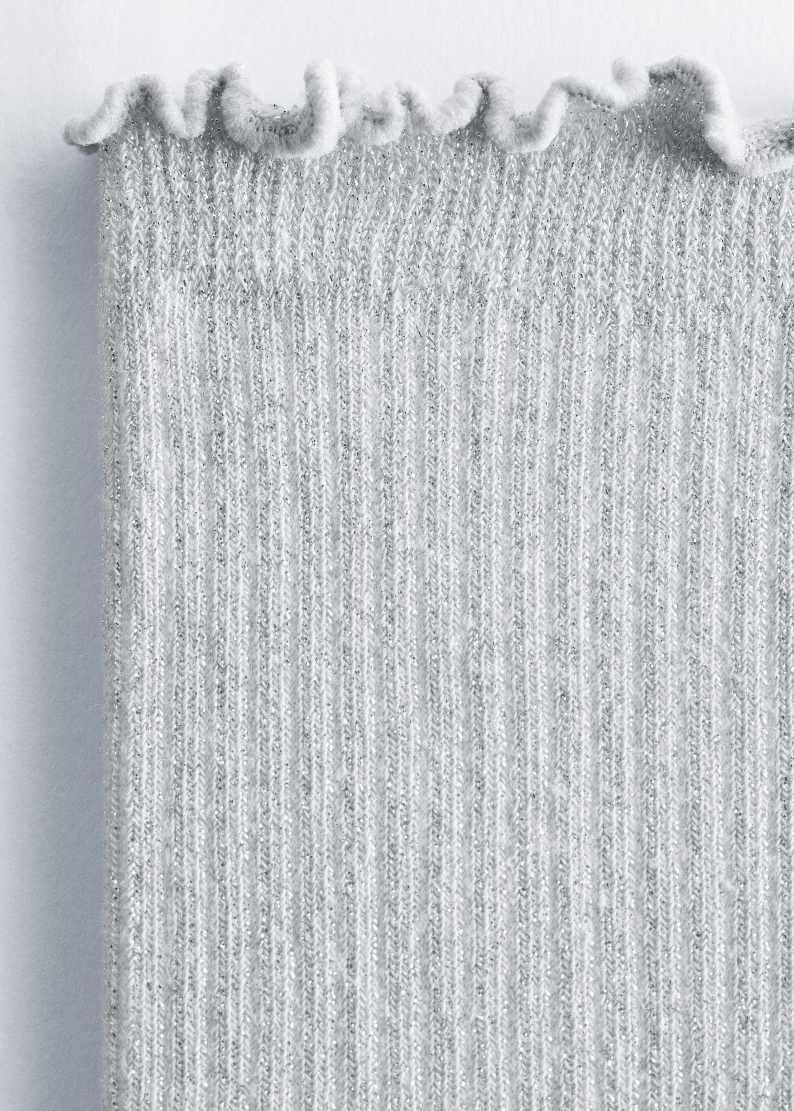 A light gray ribbed sock with a glitter lurex finish displays a shimmering, textured surface with fine vertical ribbing. The sock features a delicate, wavy frilled cuff along the top edge.