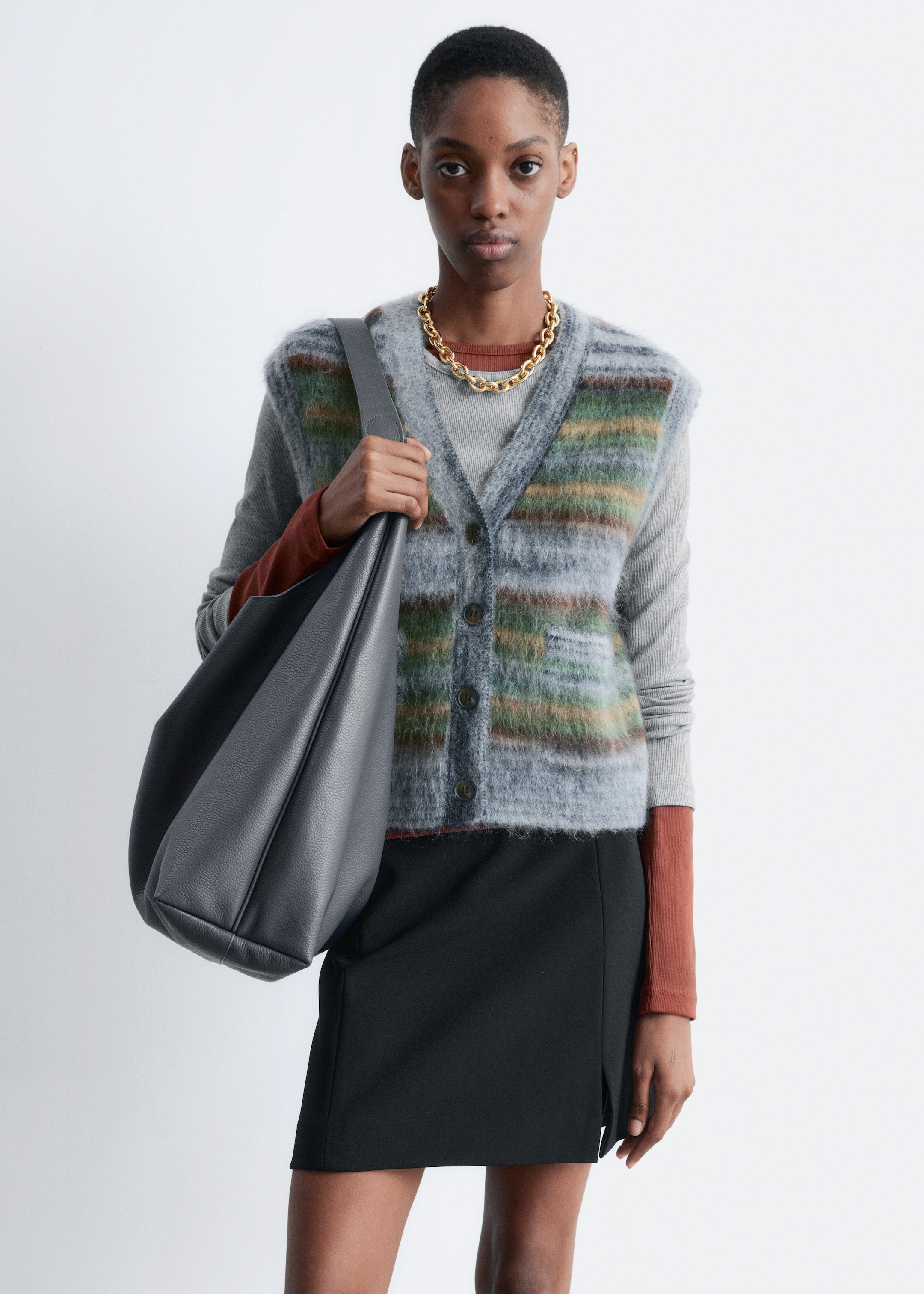 Visualizza immagine più grande: Il gilet in misto mohair, caratterizzato da un motivo a righe spazzolate in grigio, verde e marrone, è indossato dalla modella. Presenta una scollatura a V, bordi a coste, due tasche applicate sul davanti e abbottonatura lungo il davanti, ed è sovrapposto a un top a maniche lunghe e abbinato a una collana a catena dorata.