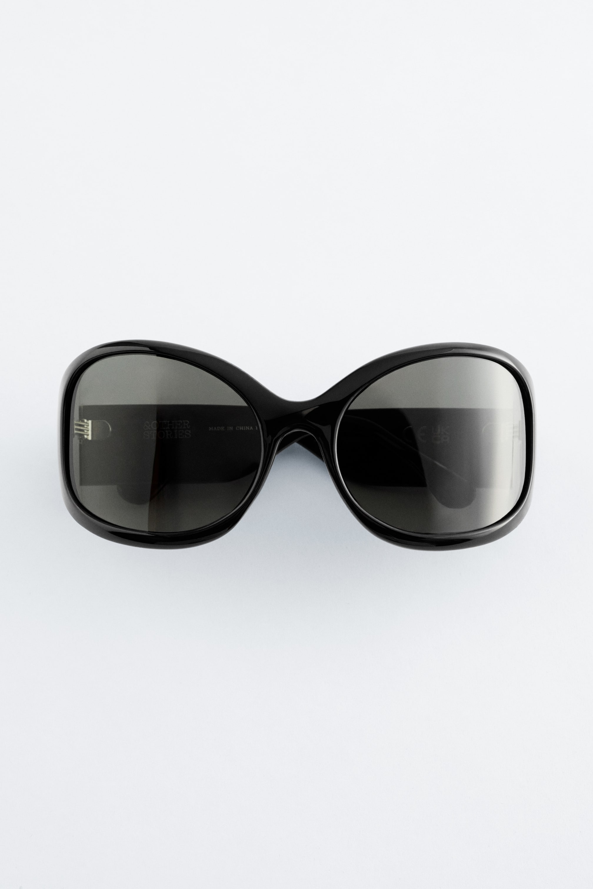 Ingrandisci l'immagine: Un paio di occhiali da sole oversize è rivolto frontalmente, caratterizzato da una montatura nera, dolcemente arrotondata e quadrata, con lenti scure sfumate e aste larghe.