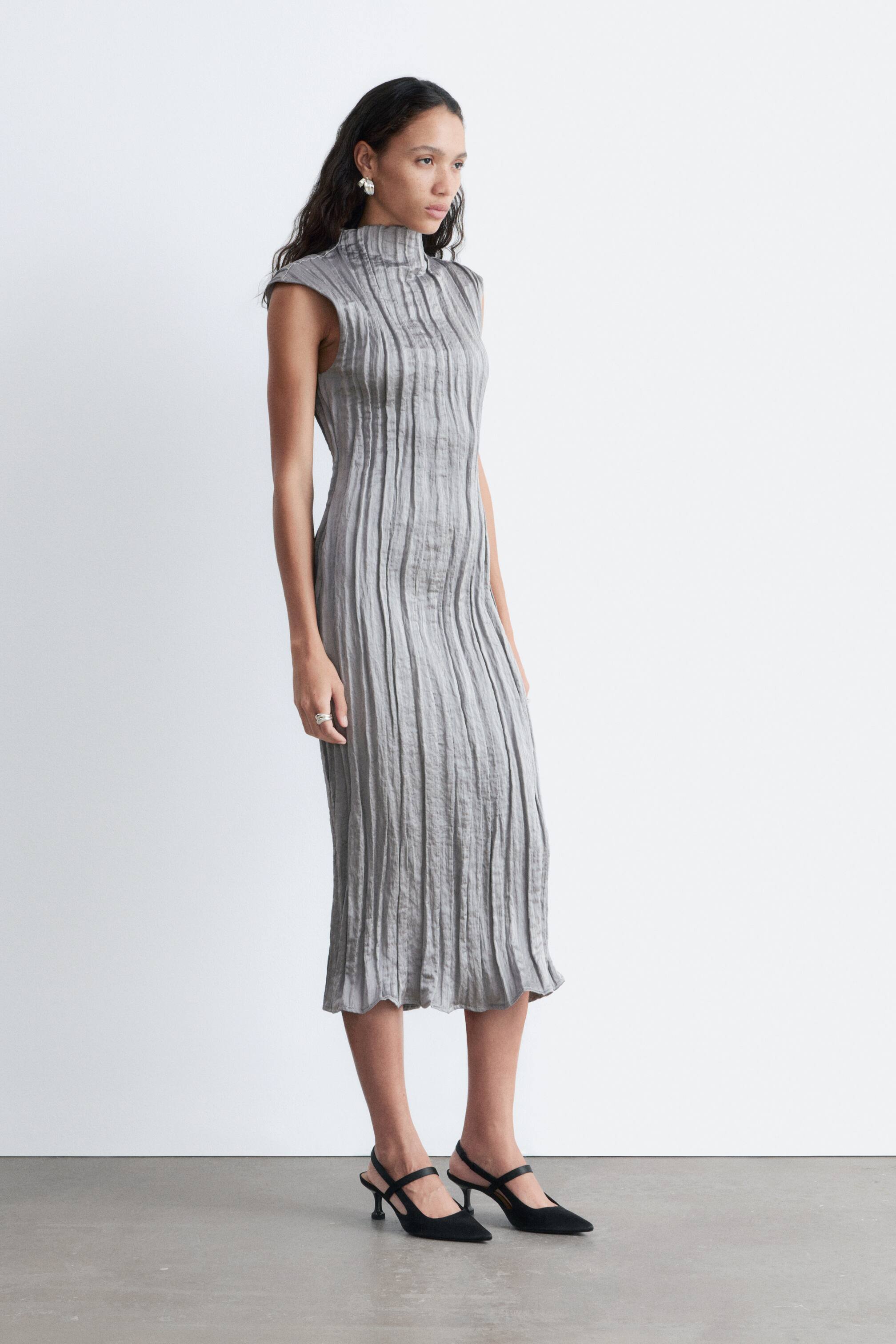 Nagyobb kép megtekintése: Egy szürke, ujjatlan midi ruha, melyet állógallérral láttak el, és végig függőleges ráncolt redőzéssel van kialakítva, enyhén oldalról bemutatva.