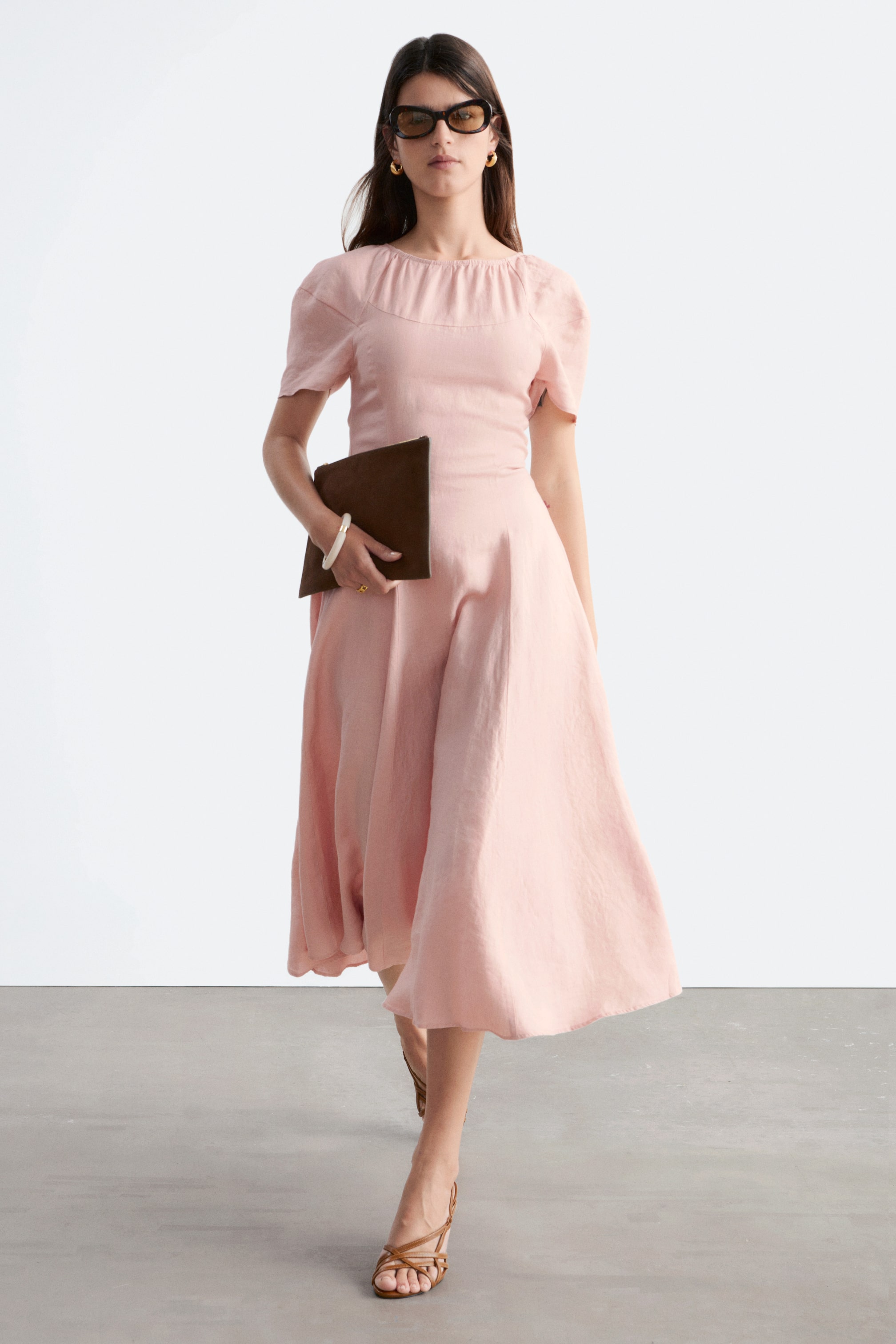 Grotere afbeelding bekijken: Een lichtroze midi-jurk in een gestructureerde stof is zichtbaar vanuit een lichte voor-zijhoek, met een geplooide ronde halslijn en korte pofmouwen. Het toont een aansluitend lijfje en een vloeiende A-lijn rok die tot halverwege de kuit reikt.