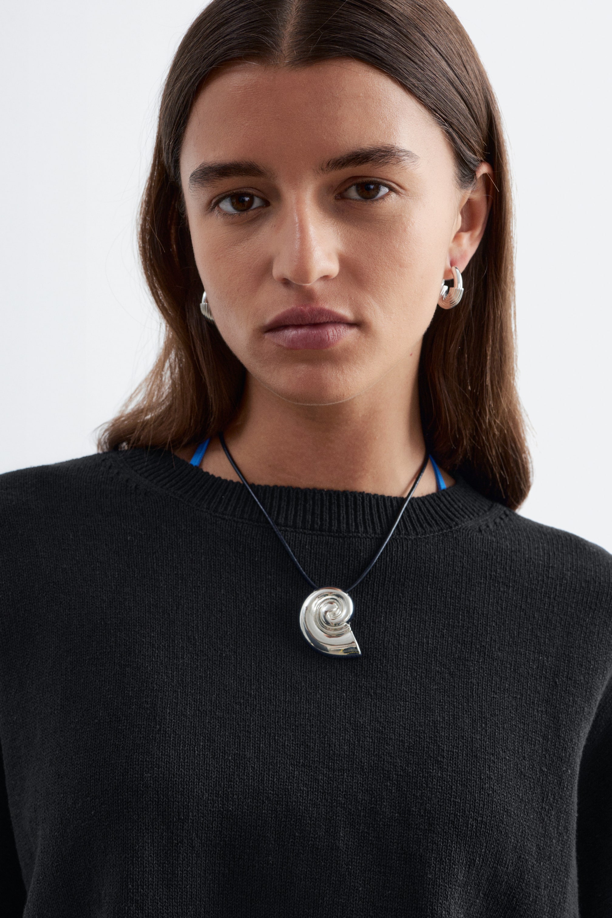 Grotere afbeelding bekijken: Een zwarte gebreide trui met een geribbelde ronde hals wordt gedragen door het model. Een zilveren spiraalhanger aan een zwarte koordketting rust op de trui. Een blauwe band is ook zichtbaar onder de halslijn.