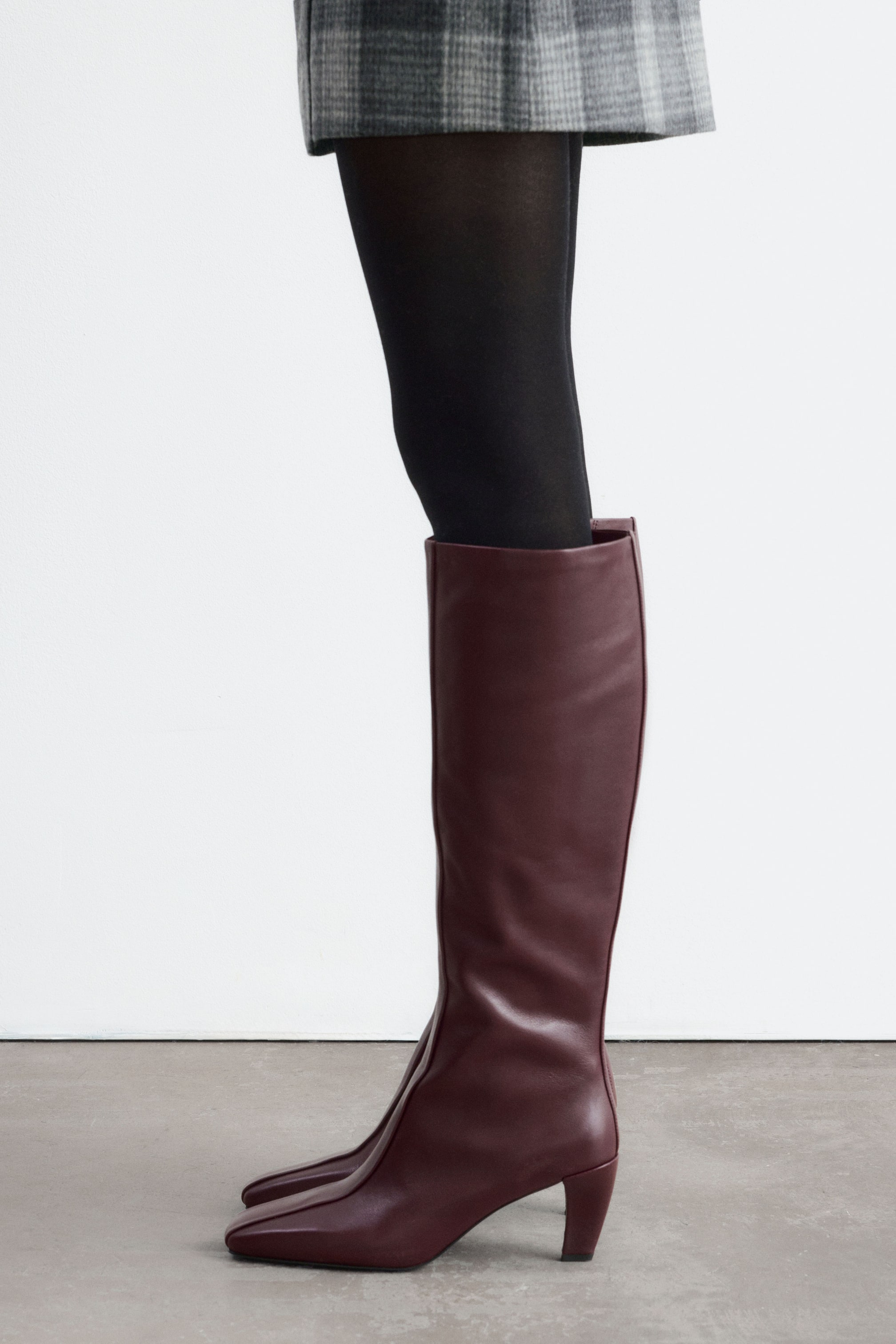 Agrandir l'image: Des bottes montantes en cuir bordeaux, dotées d’un bout carré, d’une couture avant rehaussée et d’un talon mi-haut galbé, sont portées par le mannequin. Les bottes sont associées à des collants noirs et à une jupe grise à carreaux.