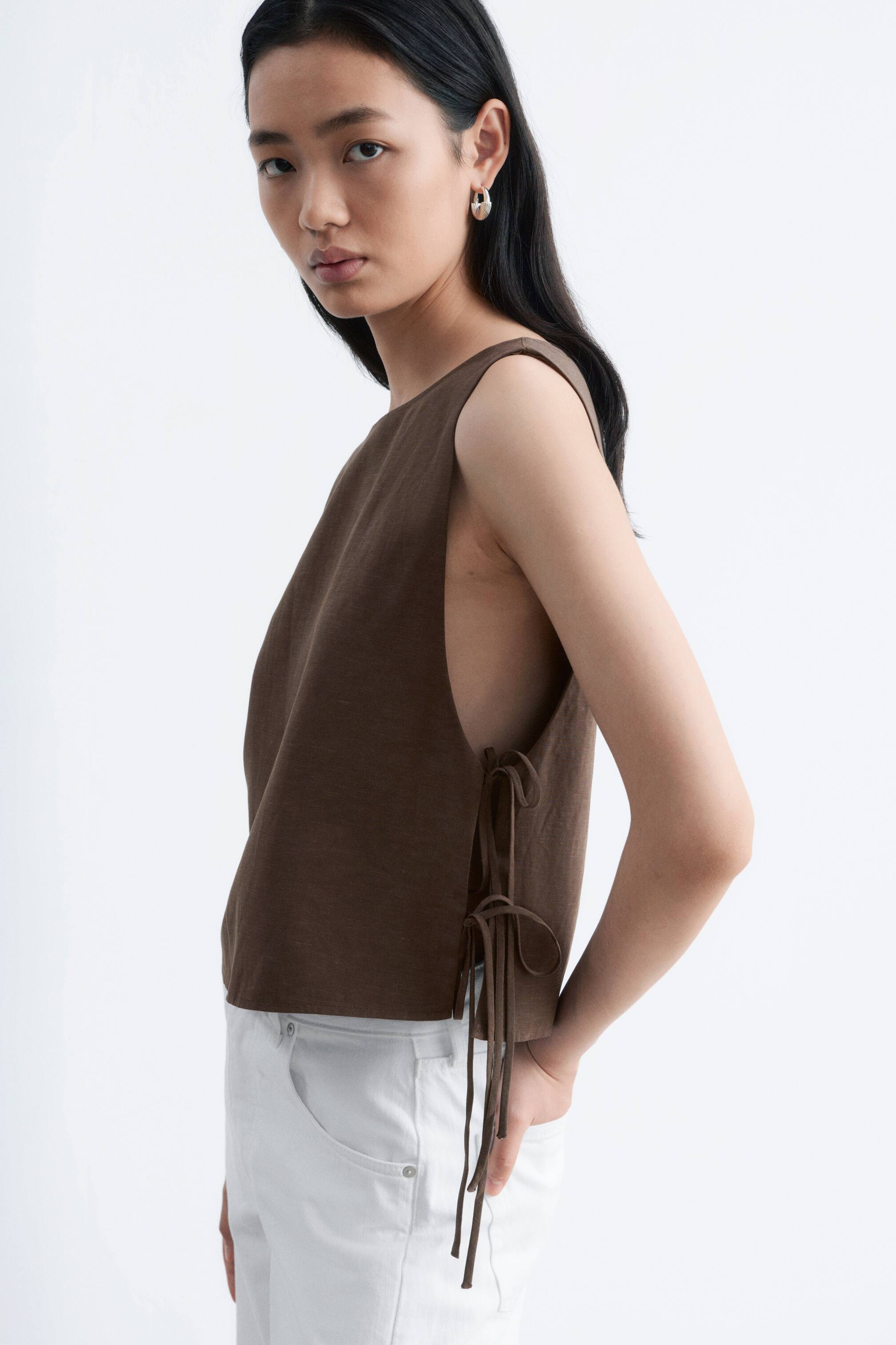 View larger image: A brown sleeveless top is being presented from a slight side angle, emphasising its right side. It features a round neck and deep armholes. Multiple fabric ties are visible on the right side, creating an adjustable and open-sided design, and the top is showing a cropped length just above the waist.