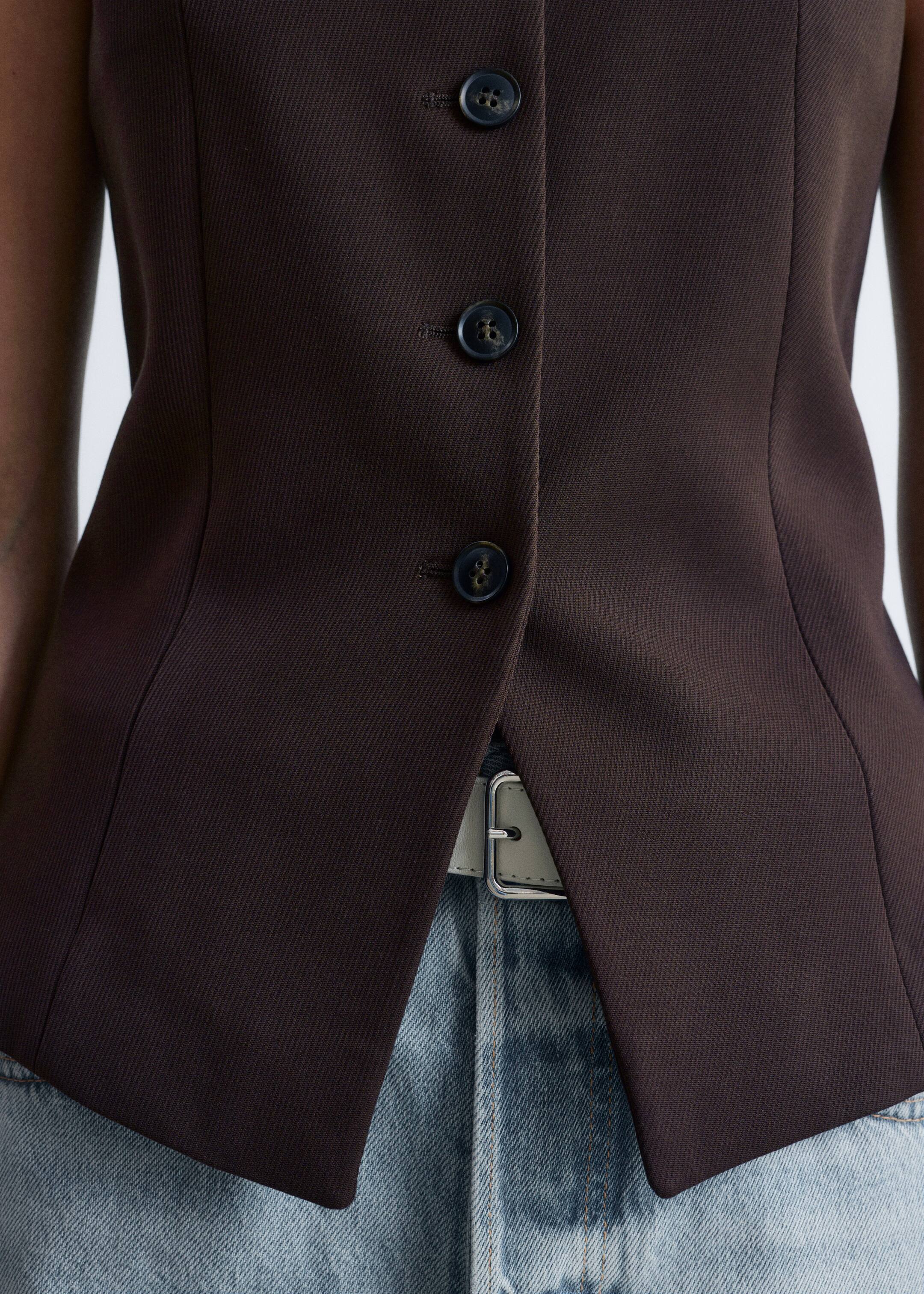 Agrandir l'image: Un gilet sans manches brun foncé est porté, avec une fermeture à trois boutons avec des boutons marbrés. Le gilet ajusté présente une couture latérale en forme, et son ourlet avant fendu révèle une ceinture de couleur claire sur un jean bleu clair.