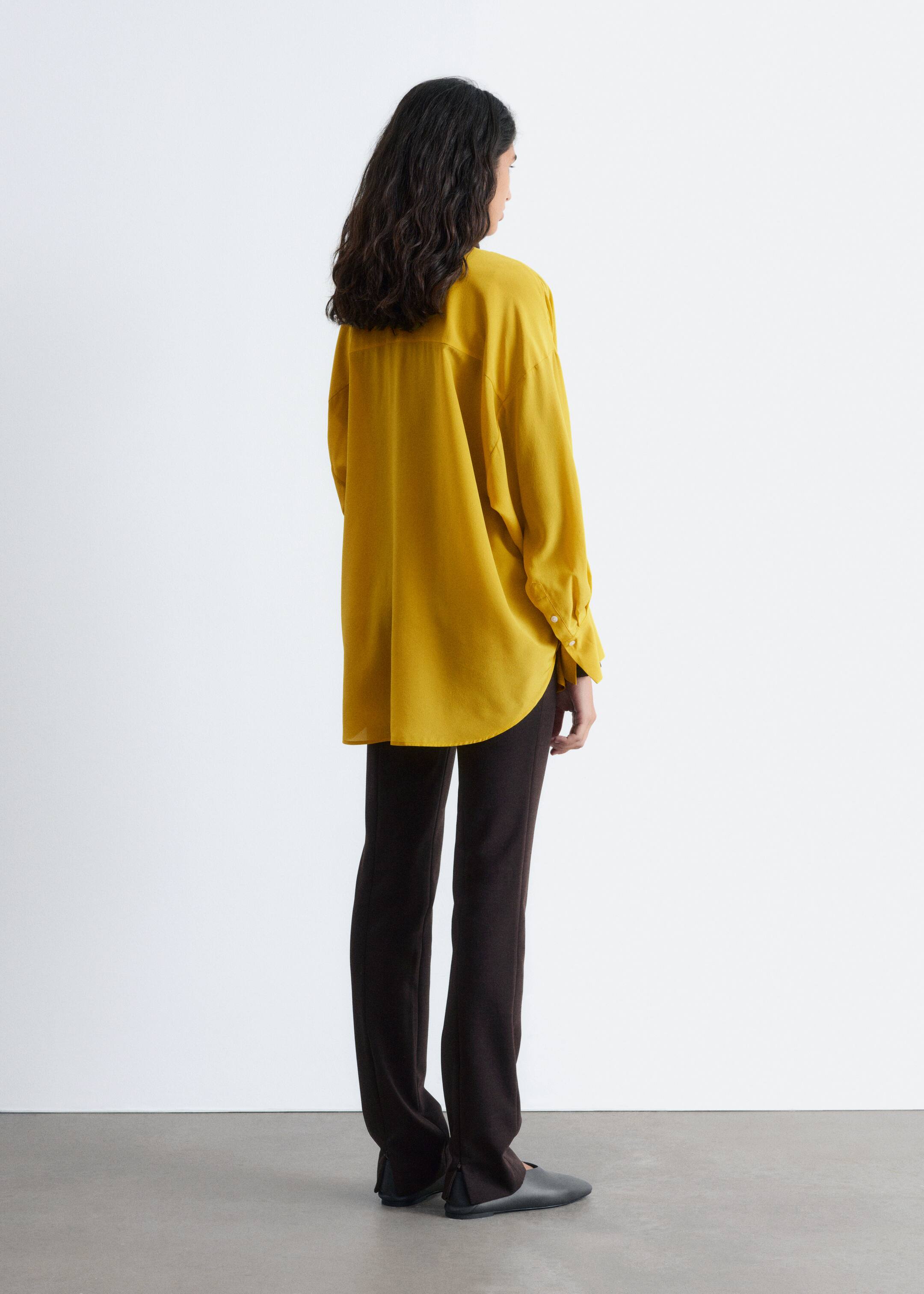 Agrandir l'image: Une chemise jaune surdimensionnée est présentée de dos et de côté, avec des manches longues à poignets boutonnés et un ourlet incurvé. Elle est stylée avec un pantalon slim-fit foncé qui a une fente subtile à la cheville, et des chaussures plates foncées complètent le look.