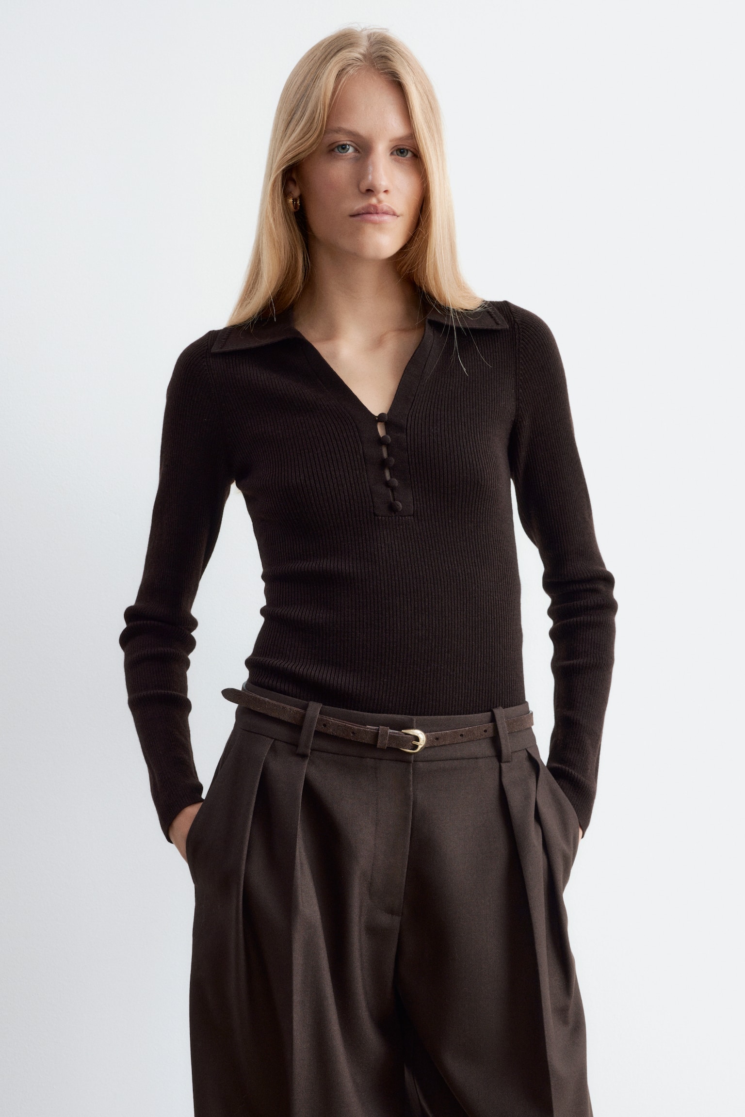 Een donkerbruine, geribde top met lange mouwen, voorzien van een kraag en een open V-hals met met stof beklede knopen, wordt gedragen door het model. De slim-fit top is ingestopt in donkerbruine geplooide broek, gestyled met een smalle riem. - 1