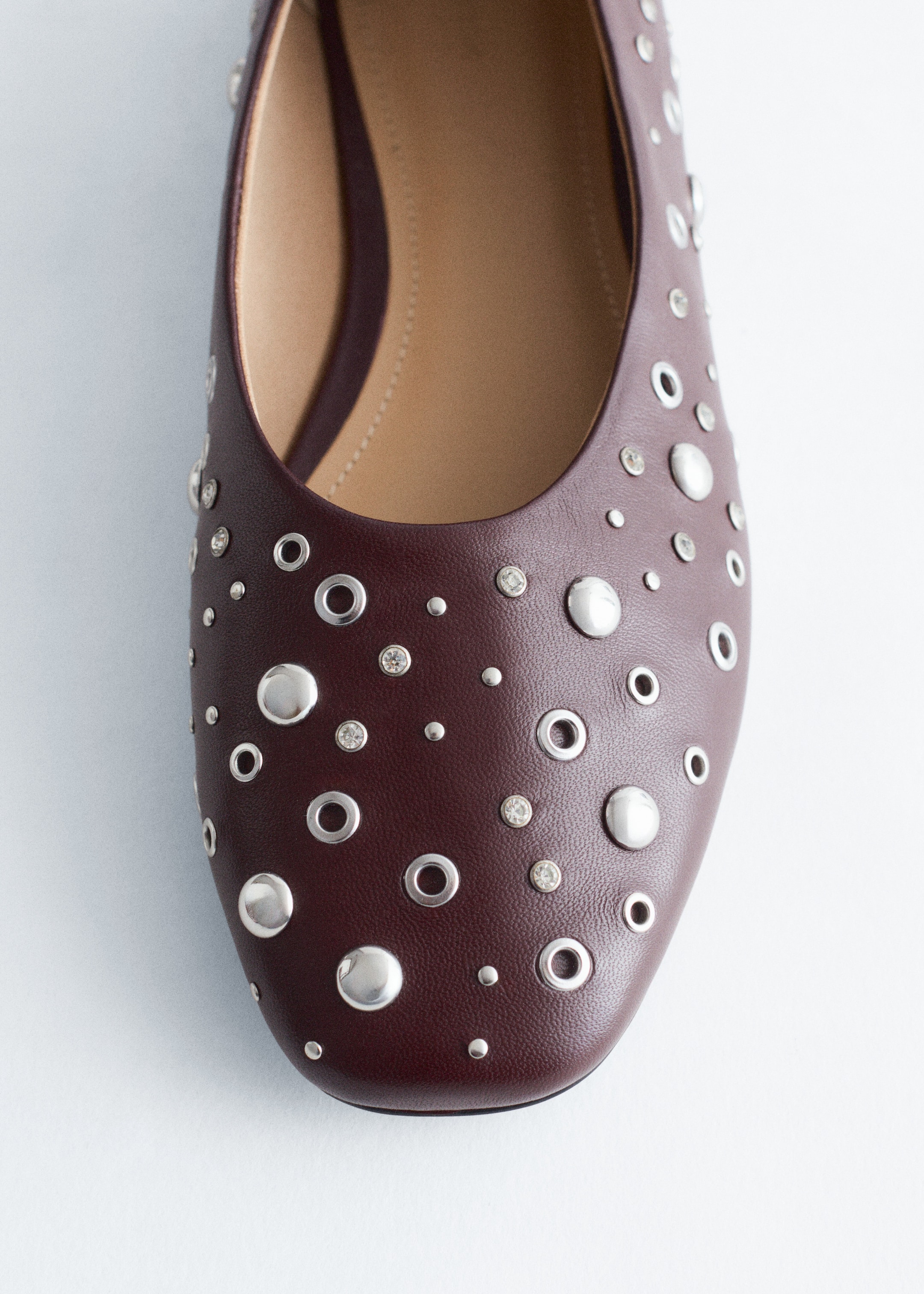Grotere afbeelding bekijken: Een bordeauxrode ballerina flat presenteert zijn voorkant, met een glad leren bovenwerk versierd met een verspreid patroon van zilverkleurige koepelnagels, kleinere ronde nagels, heldere gefacetteerde nagels en ronde oogjes. De lichtbruine binnenzool van de schoen is ook zichtbaar.