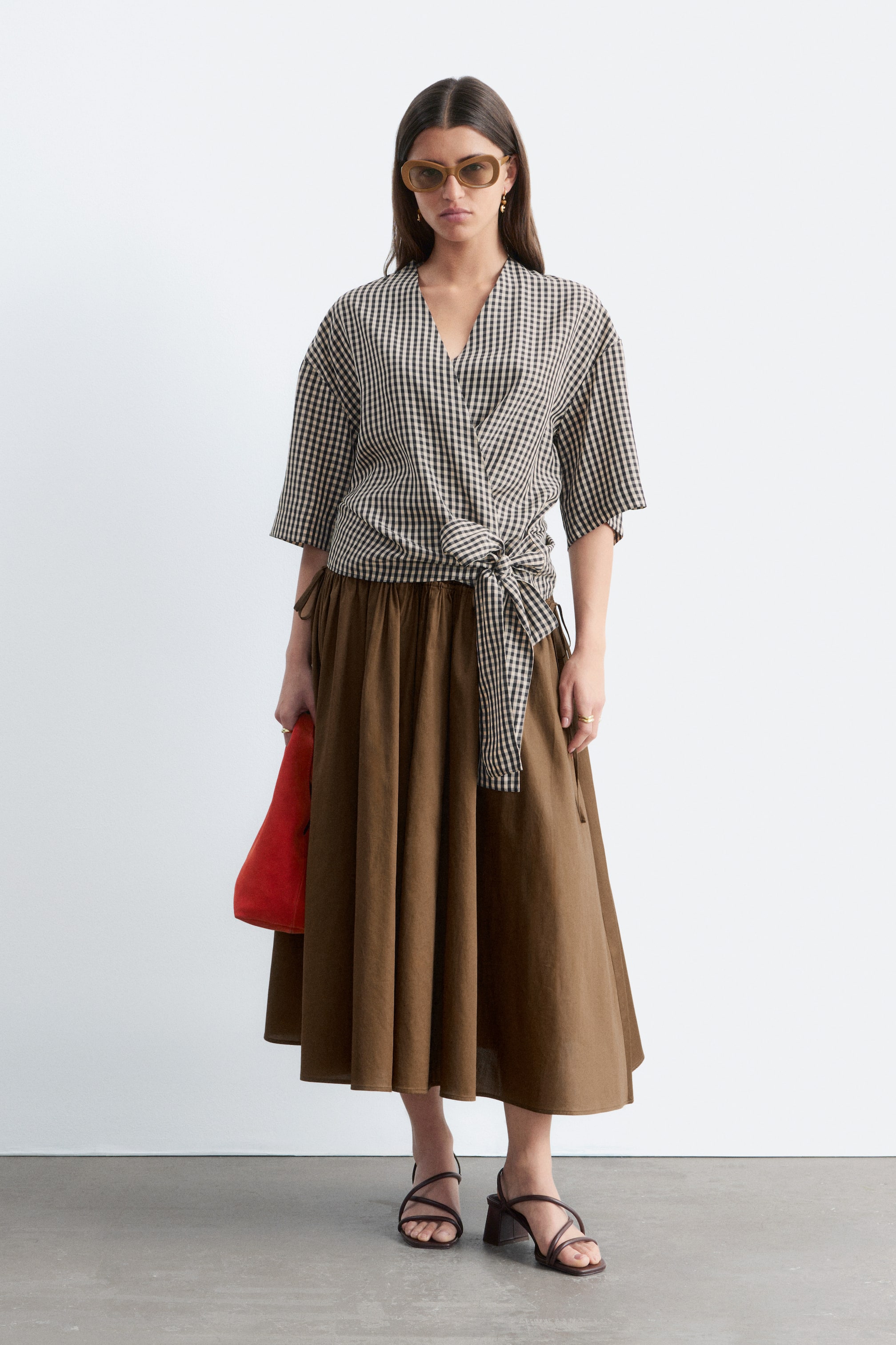 Se større billede: En sort og hvid gingham-tjekket bluse bæres af modellen, med en V-udskæring, korte ærmer og en slå-om silhuet med et selvbindende bånd, der er bundet ved fronten i taljen. Blusen er lagdelt over en brun midi-nederdel.