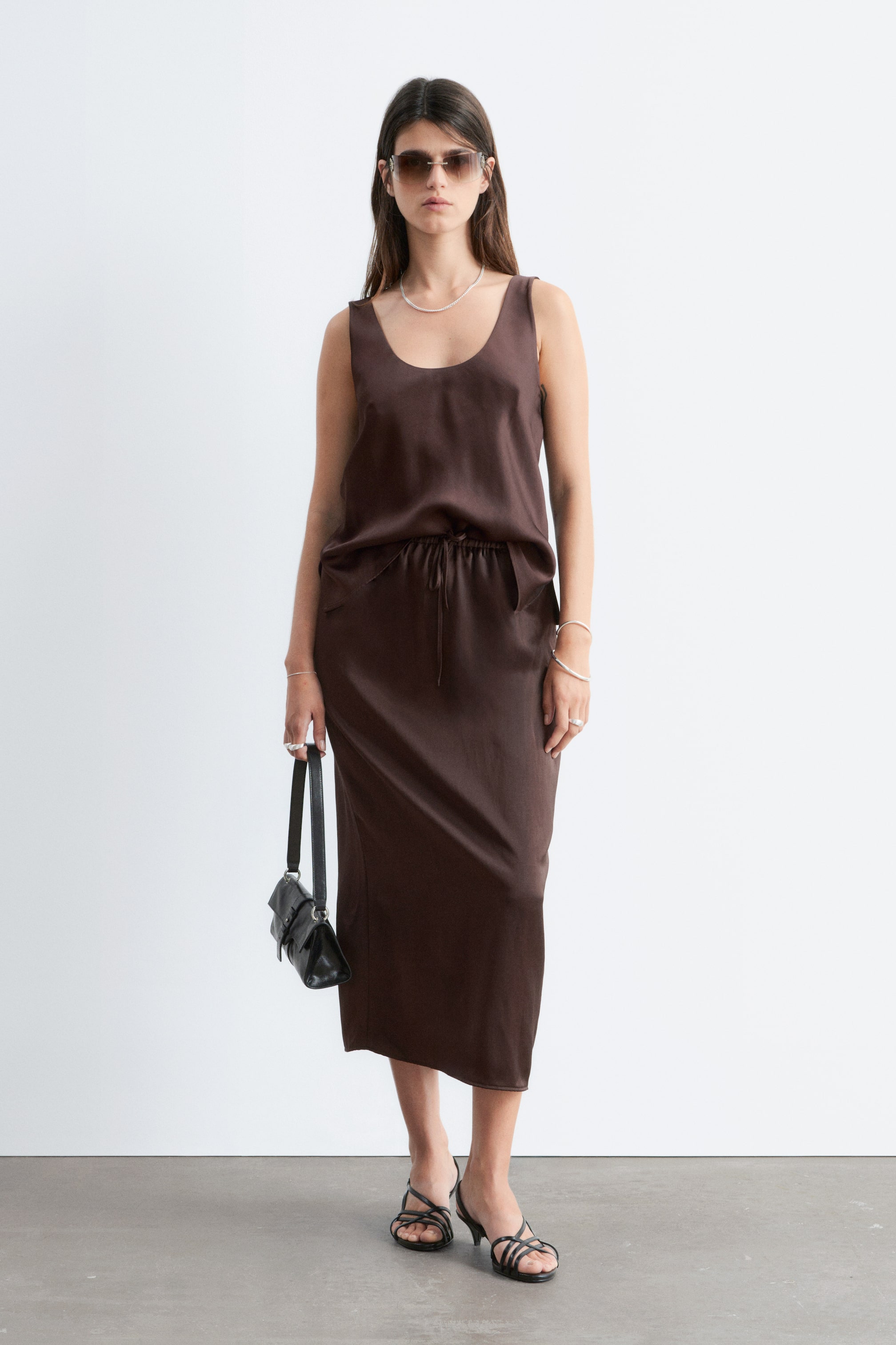 Größeres Bild anzeigen: Ein dunkelbraunes, ärmelloses Satin-Top mit Rundhalsausschnitt und einem Kordelzug, der die Taille betont, wird mit einem passenden Midi-Länge Slip-Rock präsentiert.