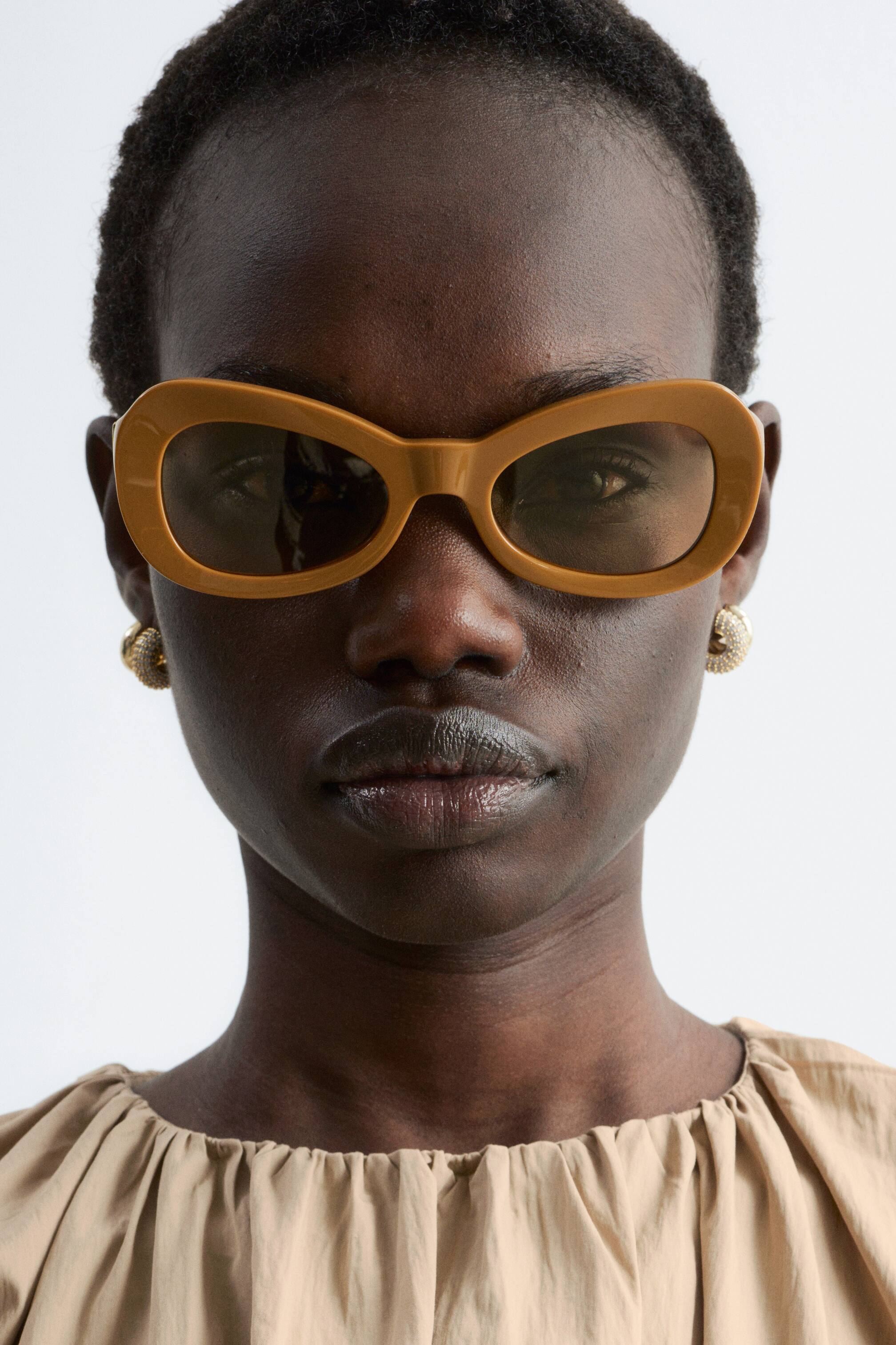 Visa större bild: Senapsgula solglasögon bärs, med tjocka, ovala bågar och mörkt tonade linser.