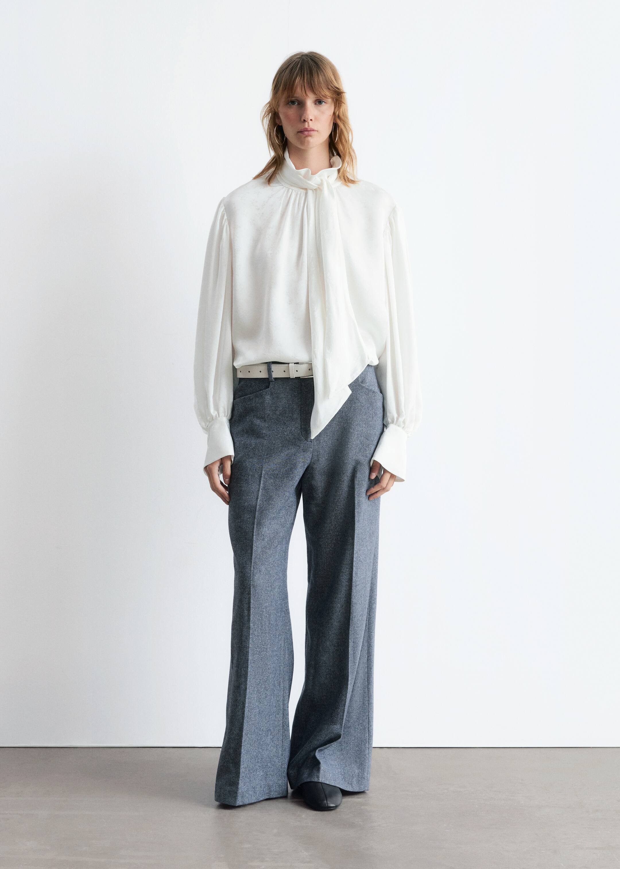 View larger image: A white blouse, made from a subtly patterned fabric, features a high stand collar and a long pussy-bow tie, with voluminous long sleeves ending in wide buttoned cuffs. It is being styled with full-length, wide-leg gray melange trousers, which are cinched with a white belt.