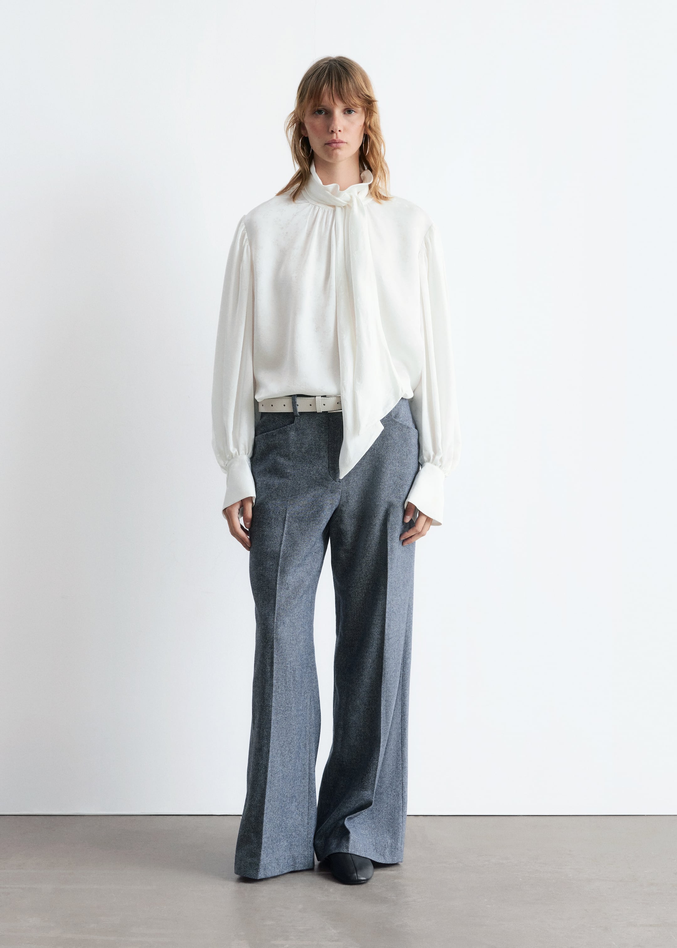Agrandir l'image: Une blouse blanche, fabriquée dans un tissu subtilement texturé, présente un col montant et un long nœud lavallière, avec des manches longues volumineuses se terminant par de larges poignets boutonnés. Elle est assortie à un pantalon gris mélangé à jambes larges et longueur complète, ceinturé d'une ceinture blanche.