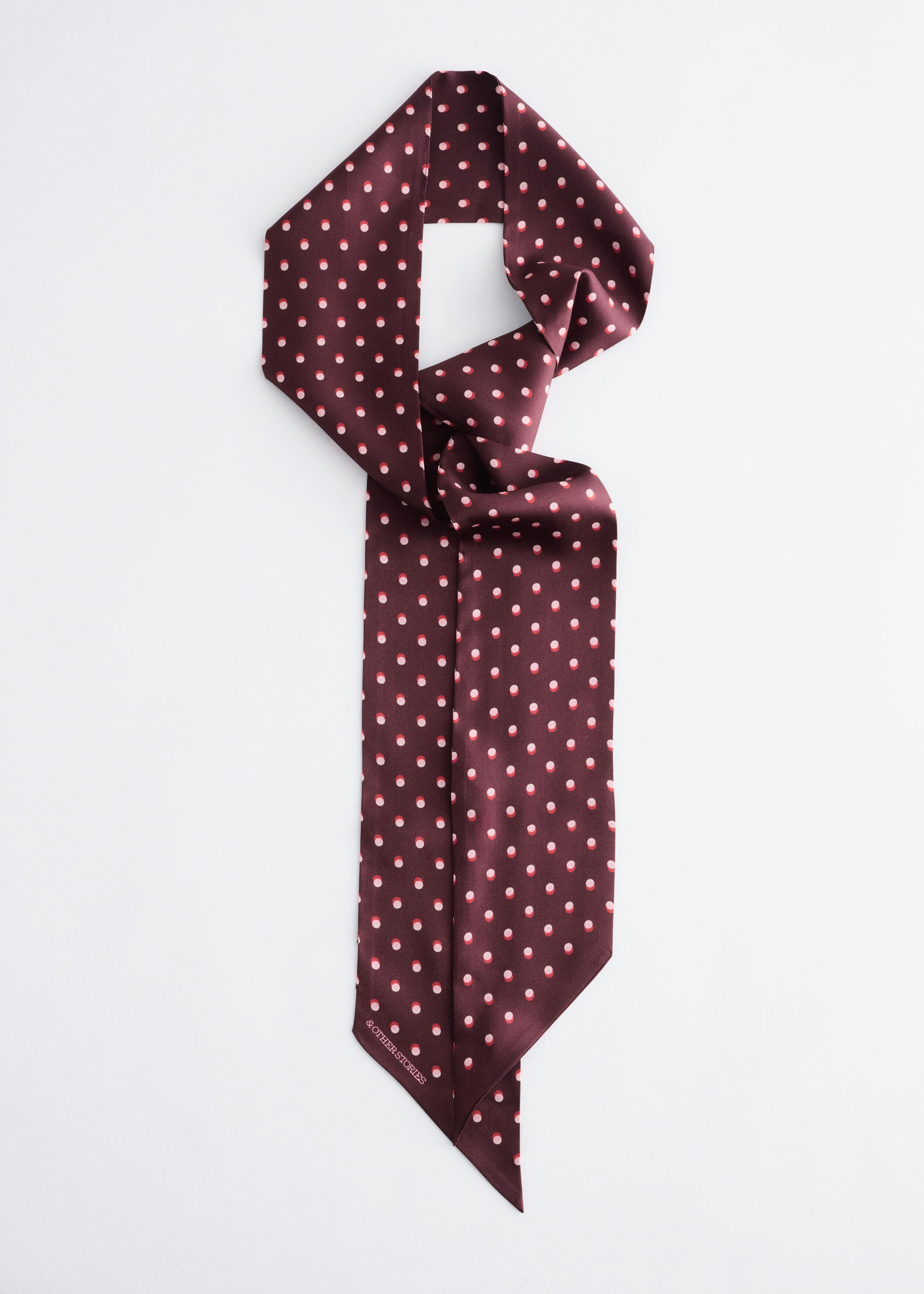 A slim, burgundy silk scarf featuring an all-over pattern of small pink and white polka dots is shown from the front. The scarf is arranged in a loose knot at the top, with its long ends hanging straight down to showcase their diagonal, pointed tips and a small logo near the bottom corner.