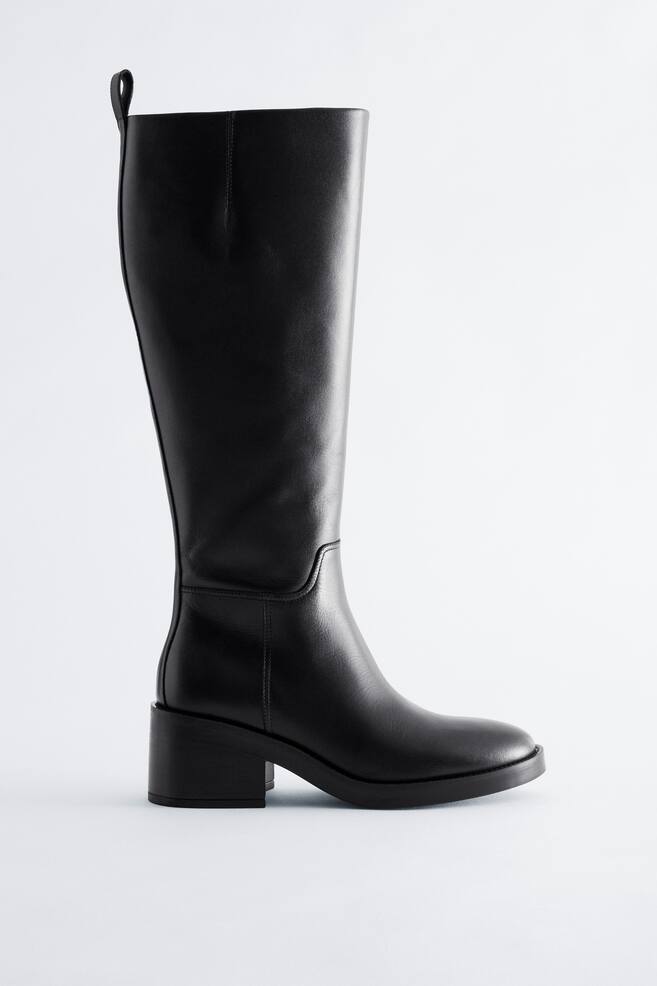 A black knee-high leather boot is shown in side profile, featuring a smooth matte finish, a rounded toe, and a low block heel. The boot features a structured shaft with a pull loop at the back collar and subtle seam detailing around the ankle. - 1