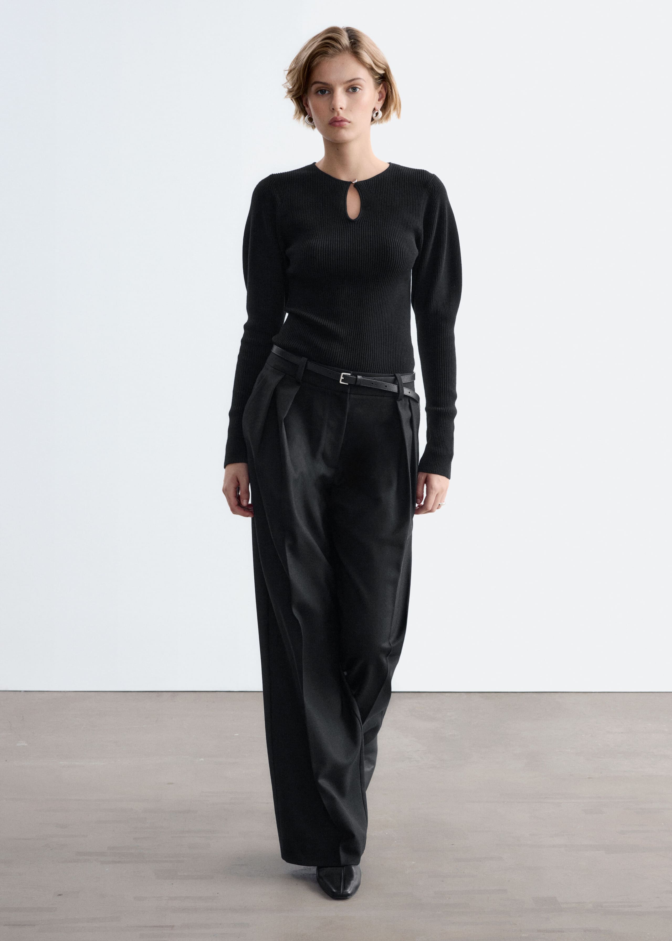 A black, rib-knit long-sleeve top with a keyhole neckline and softly puffed shoulders is worn by the model. The fitted top is tucked into high-waisted, wide-leg black trousers styled with a thin belt.