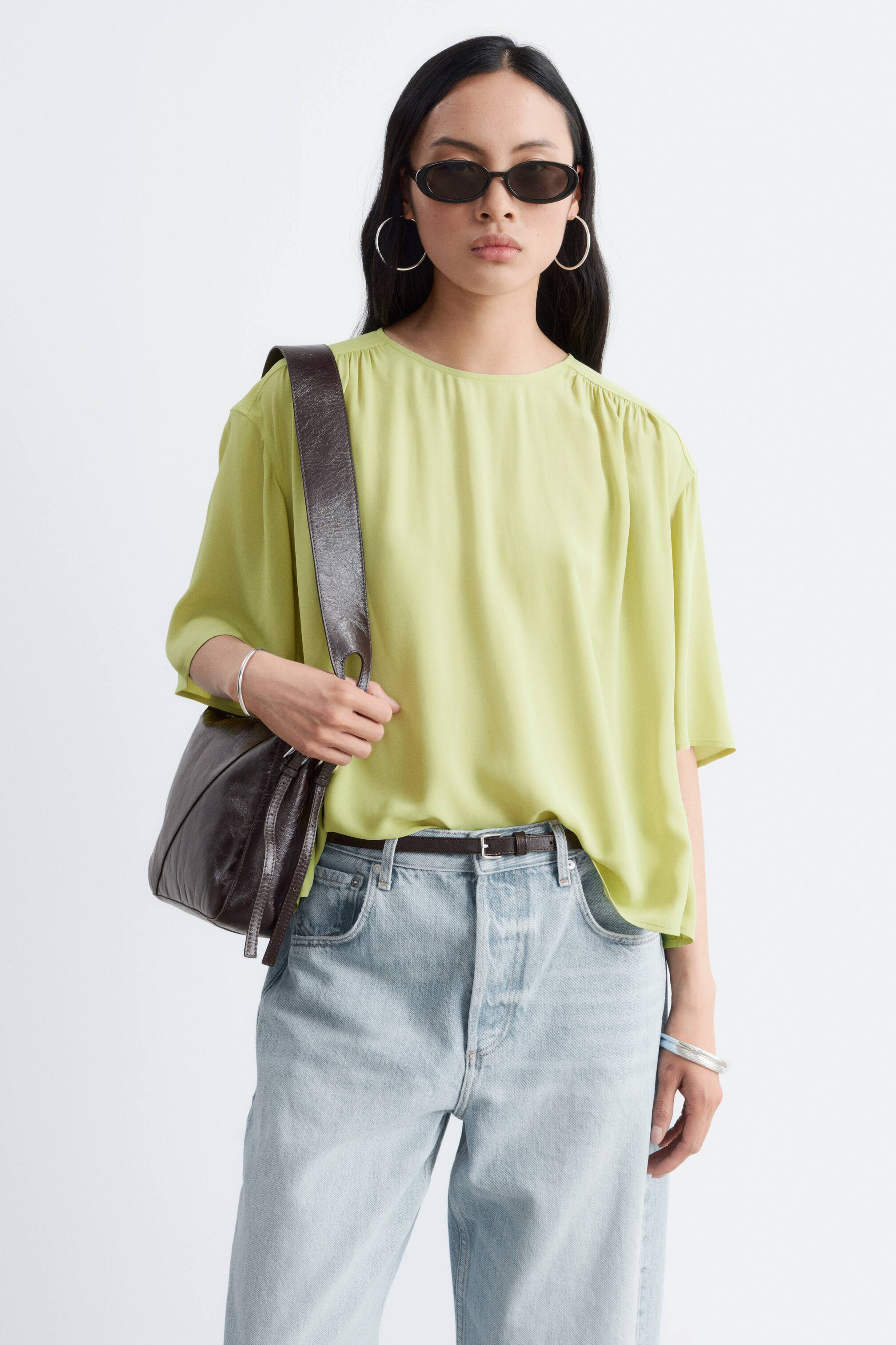 Visa större bild: En limegrön blus i flytande crepe med rund hals, korta ärmar och en överdimensionerad silhuett bärs av modellen. Den har mjuka rynkor som skapar ett mjukt, taktilt fall och är stylad instoppad i ljusa jeans med ett mörkt bälte.