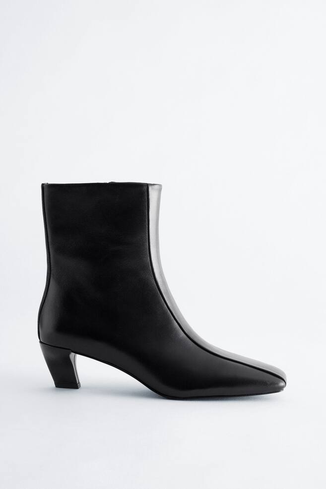 A black leather ankle boot is shown in side profile, showcasing a square toe, a low, curved kitten heel, and a central seam running down the front. - 1