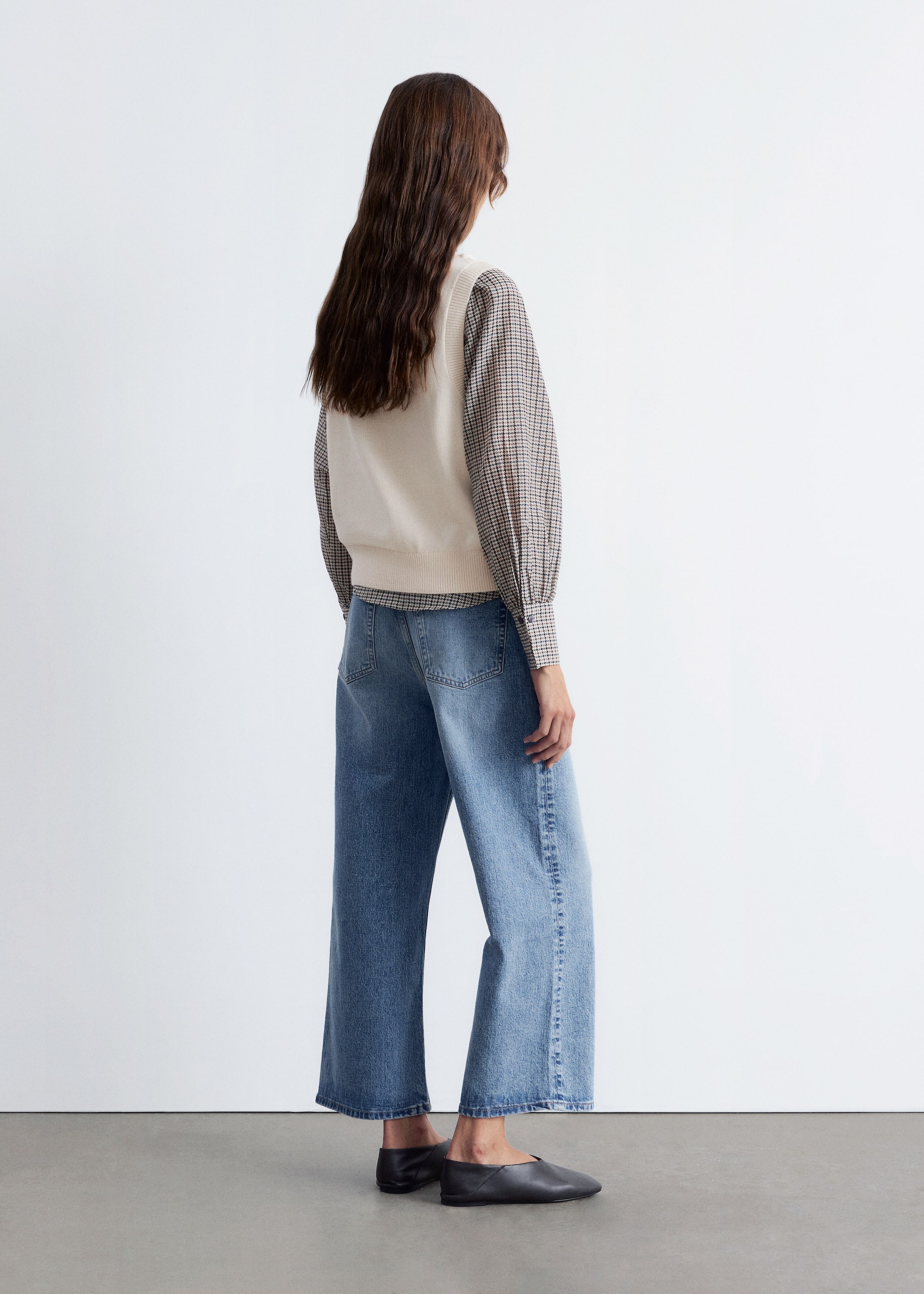 Grotere afbeelding bekijken: Een outfit wordt gepresenteerd vanuit een achteraanzicht, iets naar links gekanteld, met een crèmekleurige gebreide vest over een bruin-wit geruite overhemd met lange mouwen. De look wordt compleet gemaakt met hoog getailleerde, wijde blauwe denim jeans, met zichtbare achterzakken en een cropped lengte, gecombineerd met zwarte slip-on flats met een puntige neus.