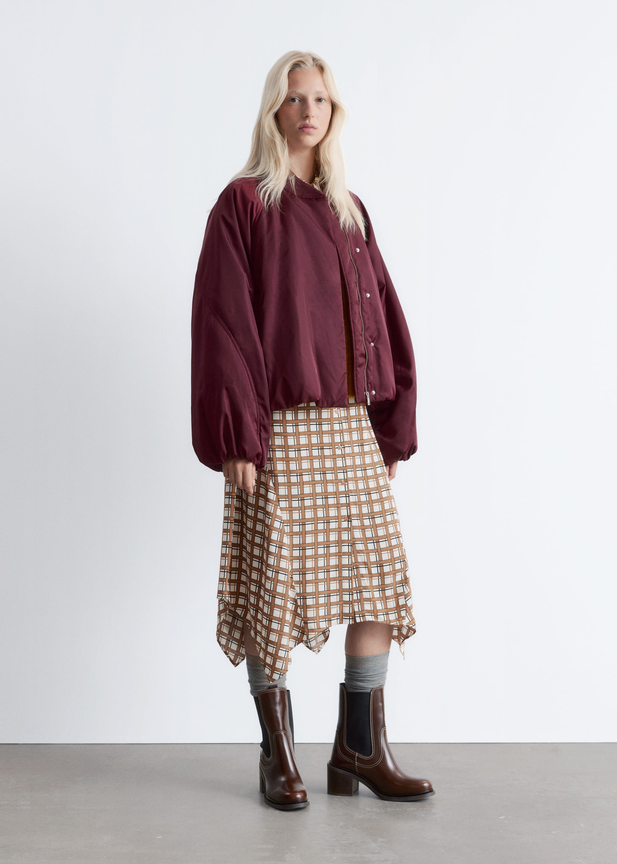 View larger image: An outfit is being presented from the front, featuring an oversized, cropped maroon bomber jacket with voluminous sleeves and an off-center snap and zipper closure over a light brown top. A brown and white plaid midi skirt with an asymmetrical handkerchief hem is being worn, paired with dark brown Chelsea boots that have a block heel and visible white stitching, and grey crew socks are showing.