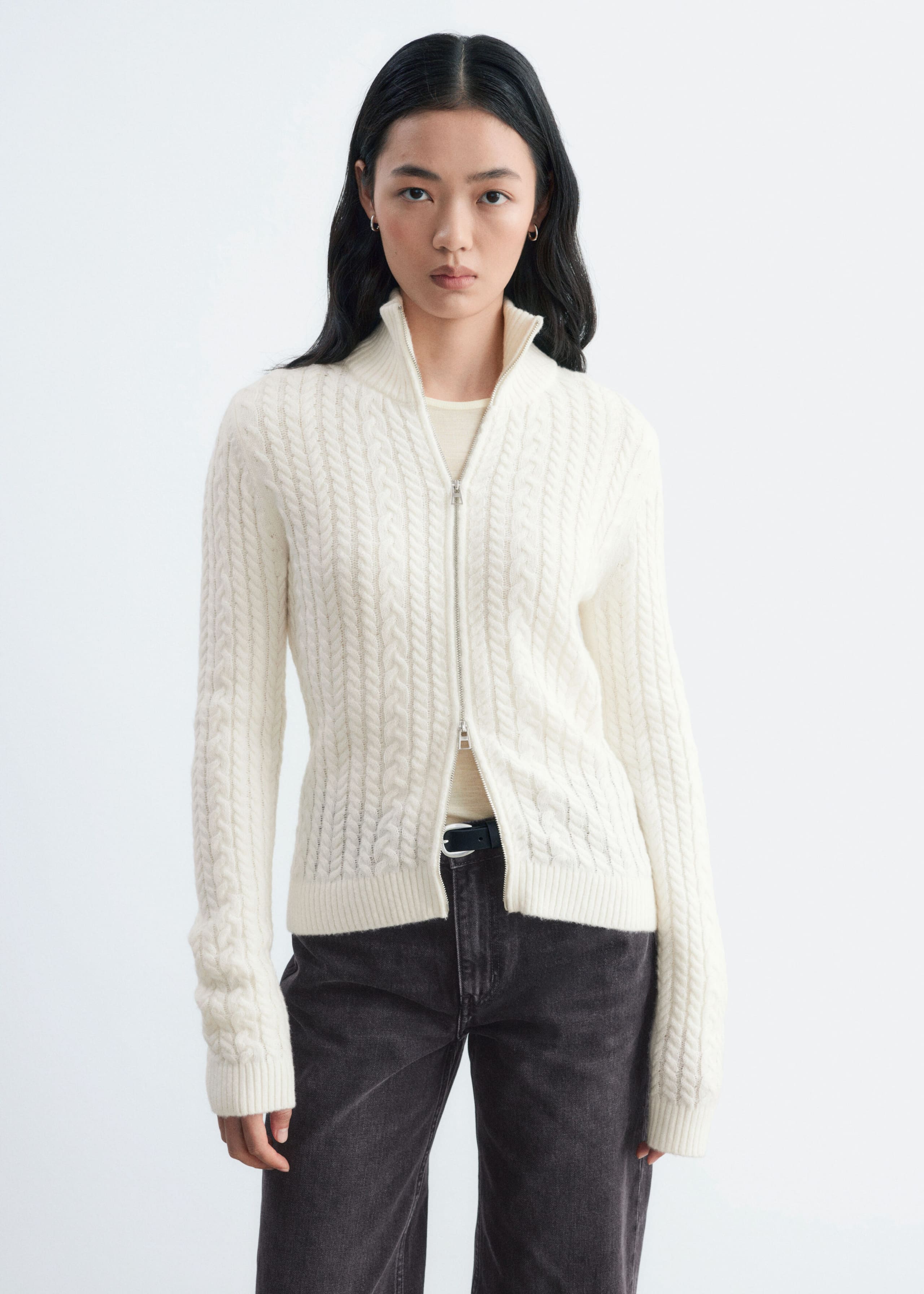 A cream-coloured, slim-fit cardigan in a mixed cable-knit pattern is being worn by the model. The cardigan features a double-layer high collar and a two-way front zip that is partially unzipped at both the top and bottom, revealing a light undershirt and dark denim jeans.