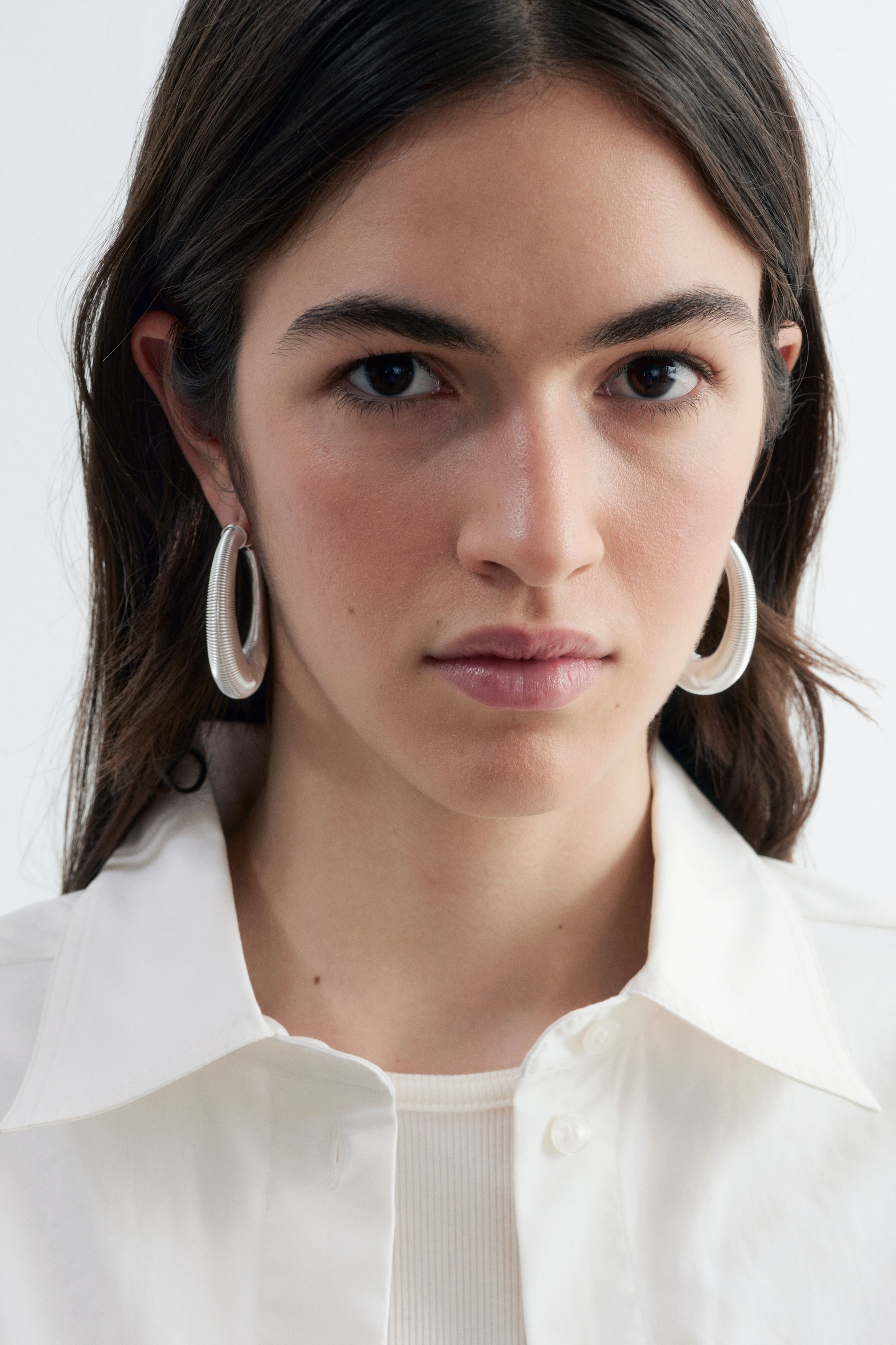 View larger image: Silver-coloured hoop earrings featuring a fine, textured ribbed finish are being worn by the model. The thick, medium-sized hoops are displayed on both ears, framed by the model's dark hair and a white collared shirt.