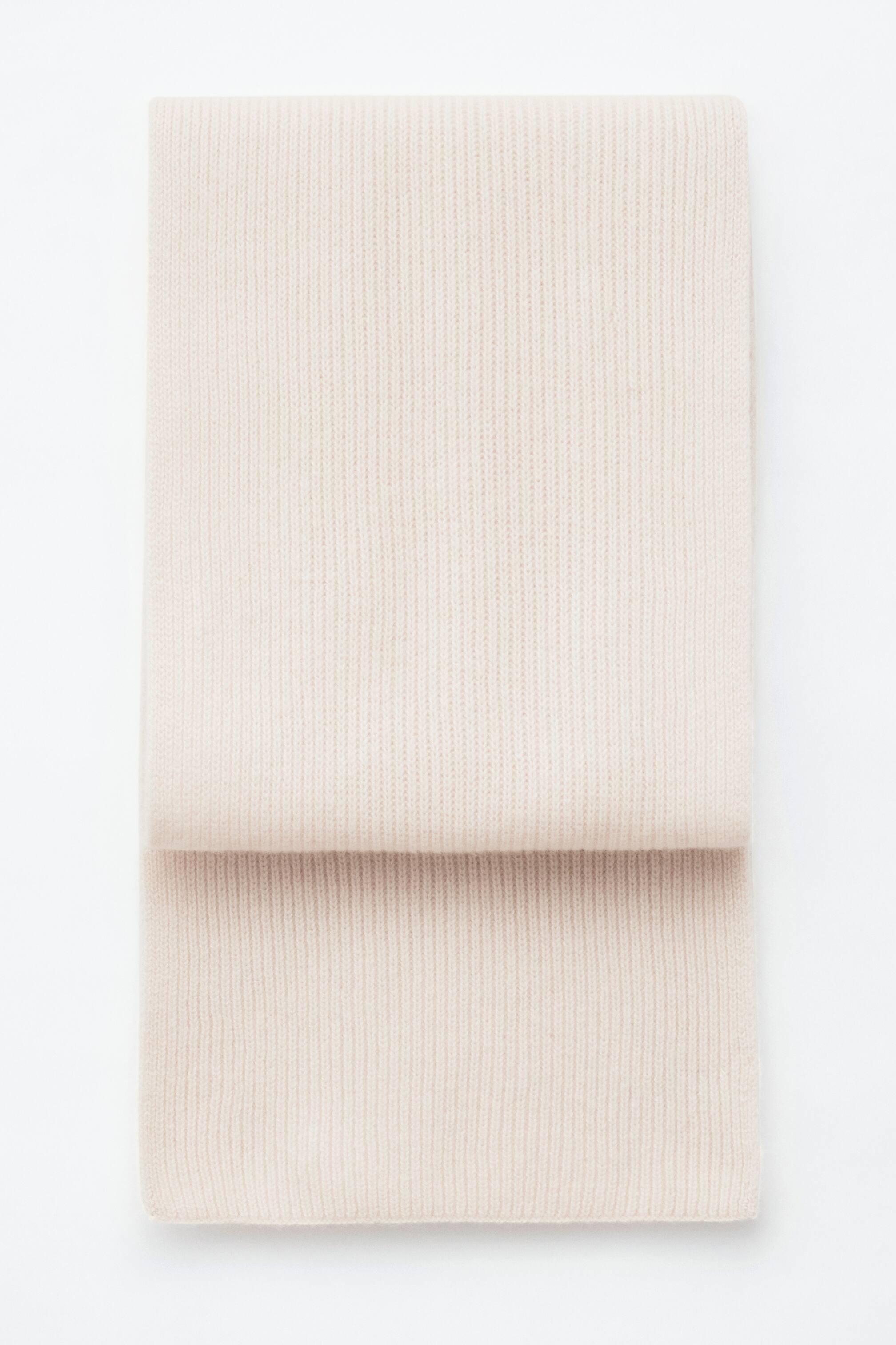 Größeres Bild anzeigen: Ein hellcremefarbener Strickschal aus Kaschmir mit vertikalem Rippenmuster liegt flach gefaltet da und zeigt seine weiche Textur sowie saubere, gerade Kanten.