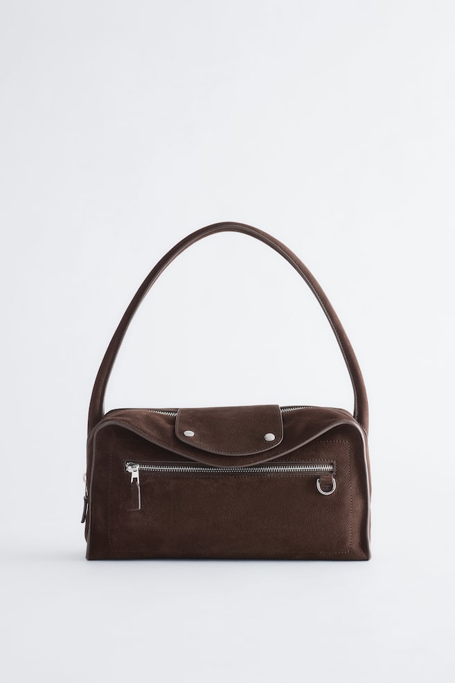 Eine dunkelbraune Schultertasche aus Wildleder steht aufrecht und zeigt einen einzelnen gebogenen Schulterriemen sowie einen rechteckigen Korpus mit geschwungener Oberlinie. Die Vorderseite präsentiert einen Ãberschlag mit zwei Druckknöpfen in Silberoptik, ein waagerechtes ReiÃverschlussfach und einen kleinen D-Ring aus Metall. - 1