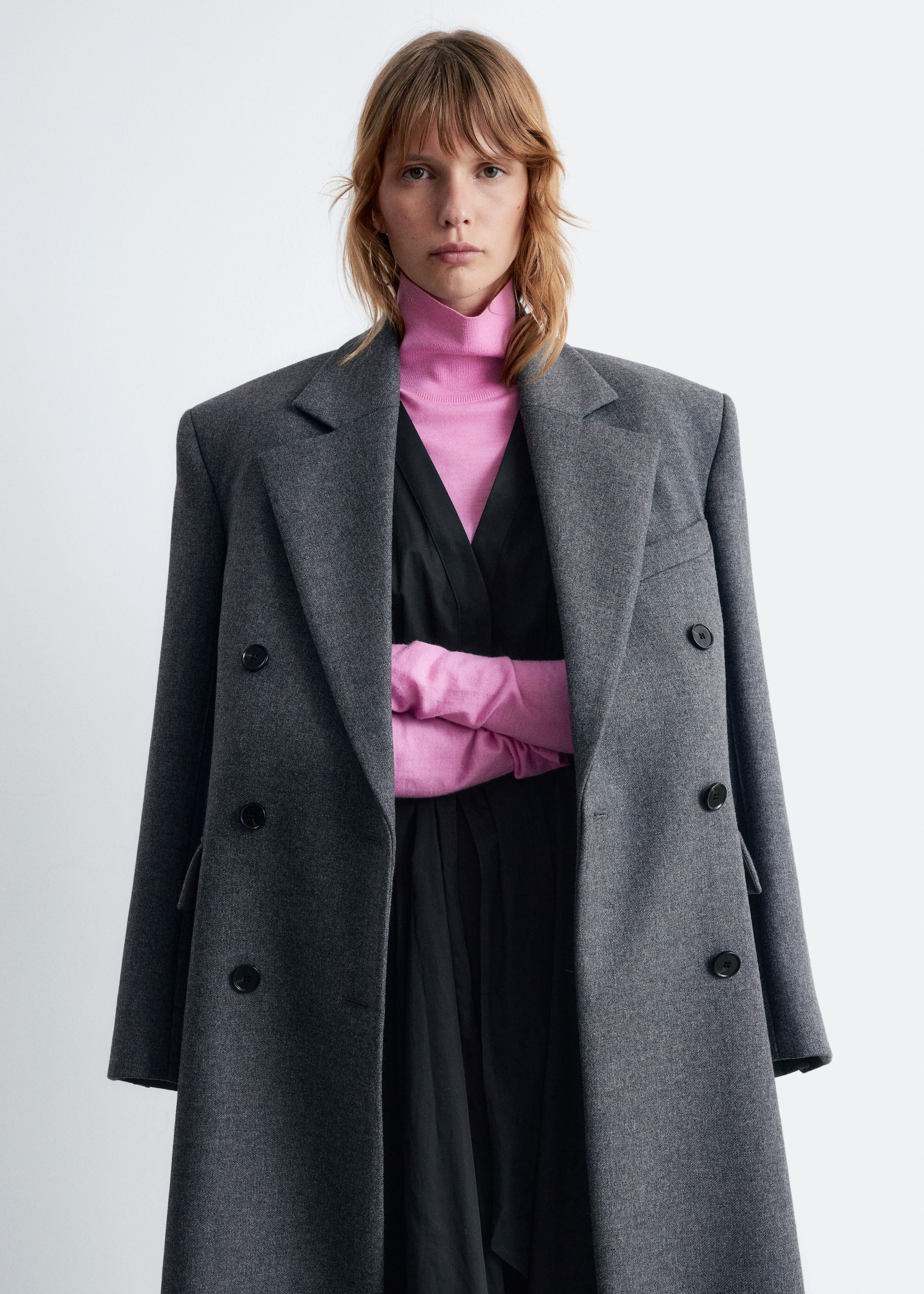 Agrandir l'image: Un manteau gris à double boutonnage est présenté de face, avec de larges revers en pointe, des épaules larges et deux boutons noirs. Il est porté par-dessus un col roulé rose et un haut noir à col en V.